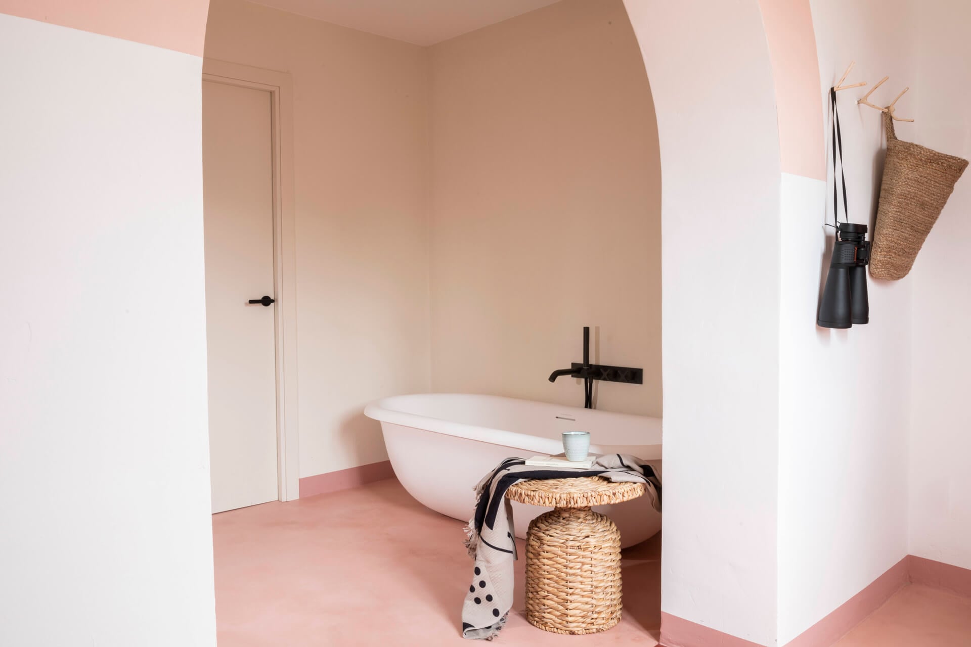 Menorca Experimental, Balearic Islands, Spain — Minimalist bathroom with curved white bathtub, terracotta floors, black fixtures, woven storage baskets, and soft pink-toned walls.