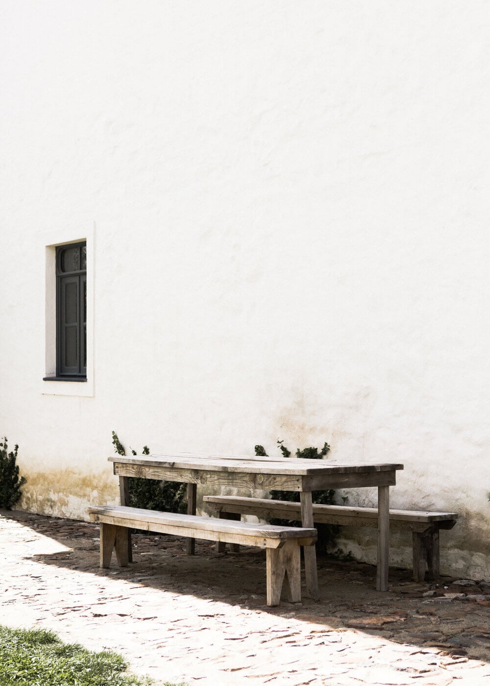 São Lourenço do Barrocal, Portugal — Weathered wooden picnic table and bench against whitewashed stone wall with dark window, rustic courtyard seating.