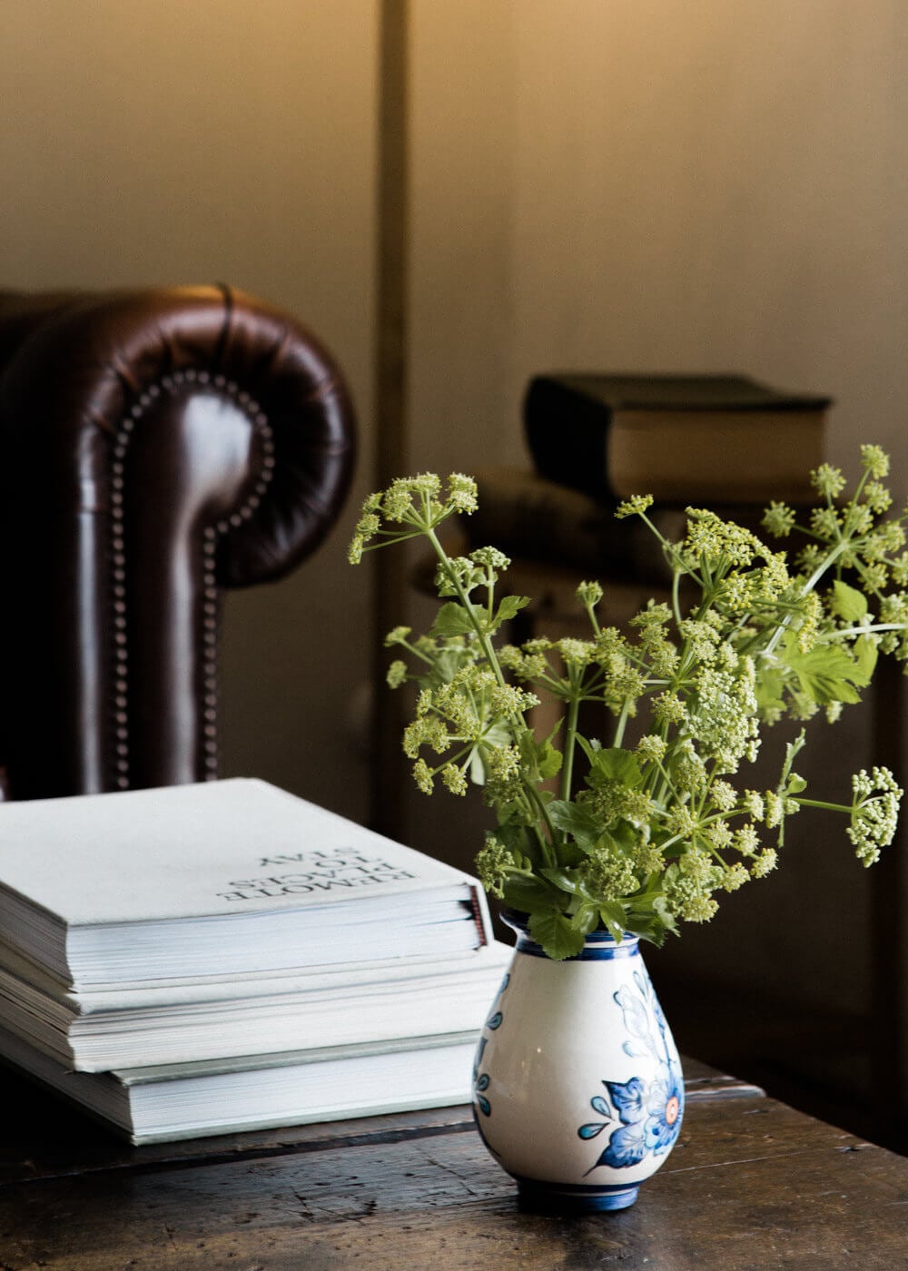 São Lourenço do Barrocal, Portugal — Stacked books beside blue and white ceramic vase with green parsley, leather wingback chair and neutral walls in refined interior.