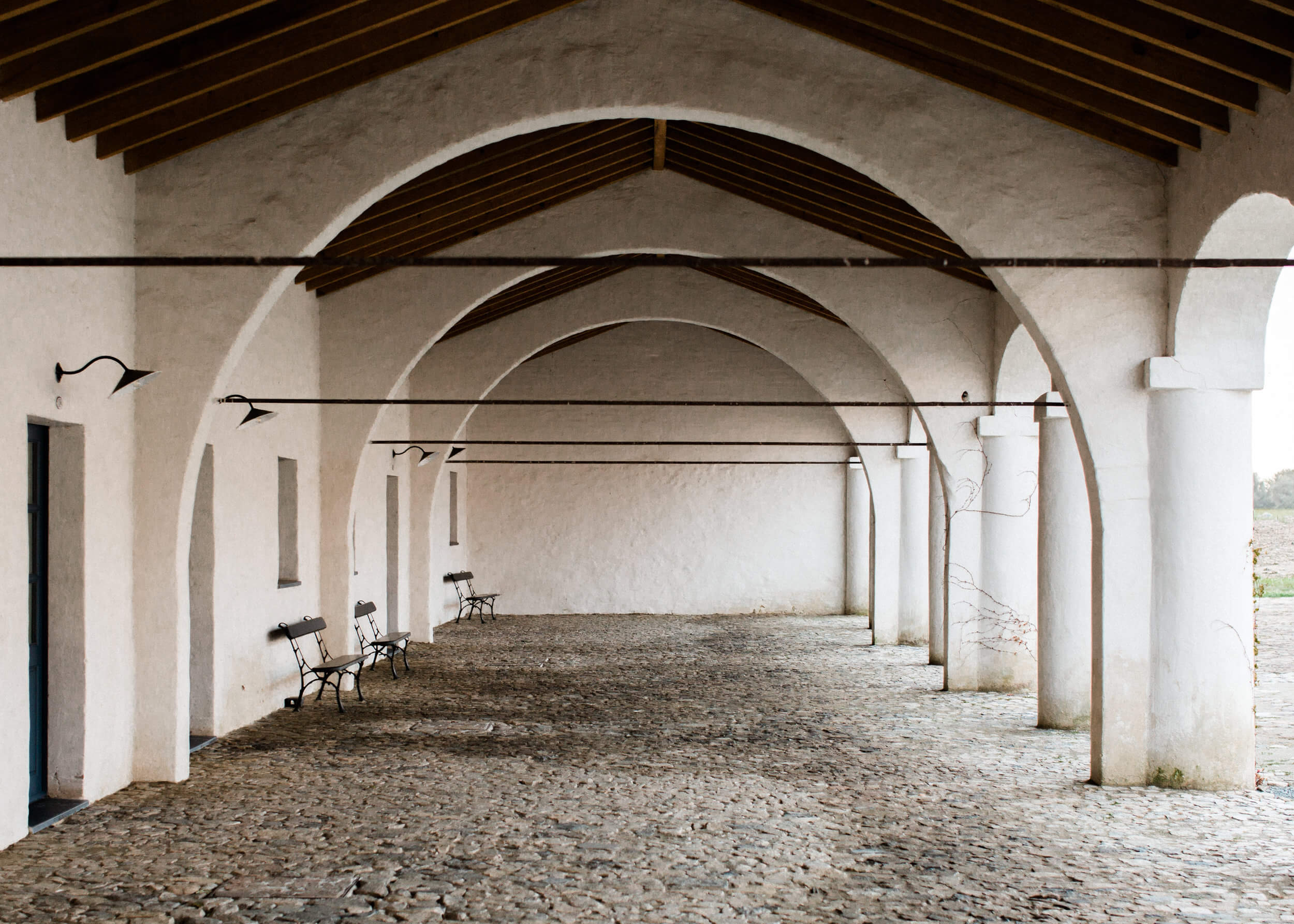 São Lourenço do Barrocal, Portugal — Arched colonnade with whitewashed walls, wooden beams, gravel floor, and iron benches in minimalist hospitality design.