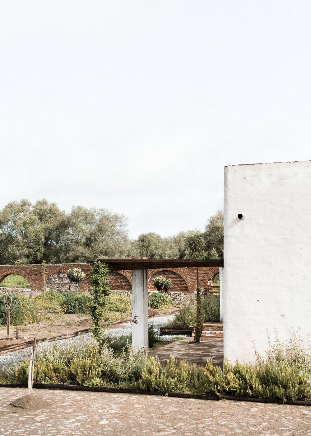 São Lourenço do Barrocal, Portugal — Modernist white tower beside terracotta courtyard with arched brick walls, olive trees, and minimalist Mediterranean landscape design.
