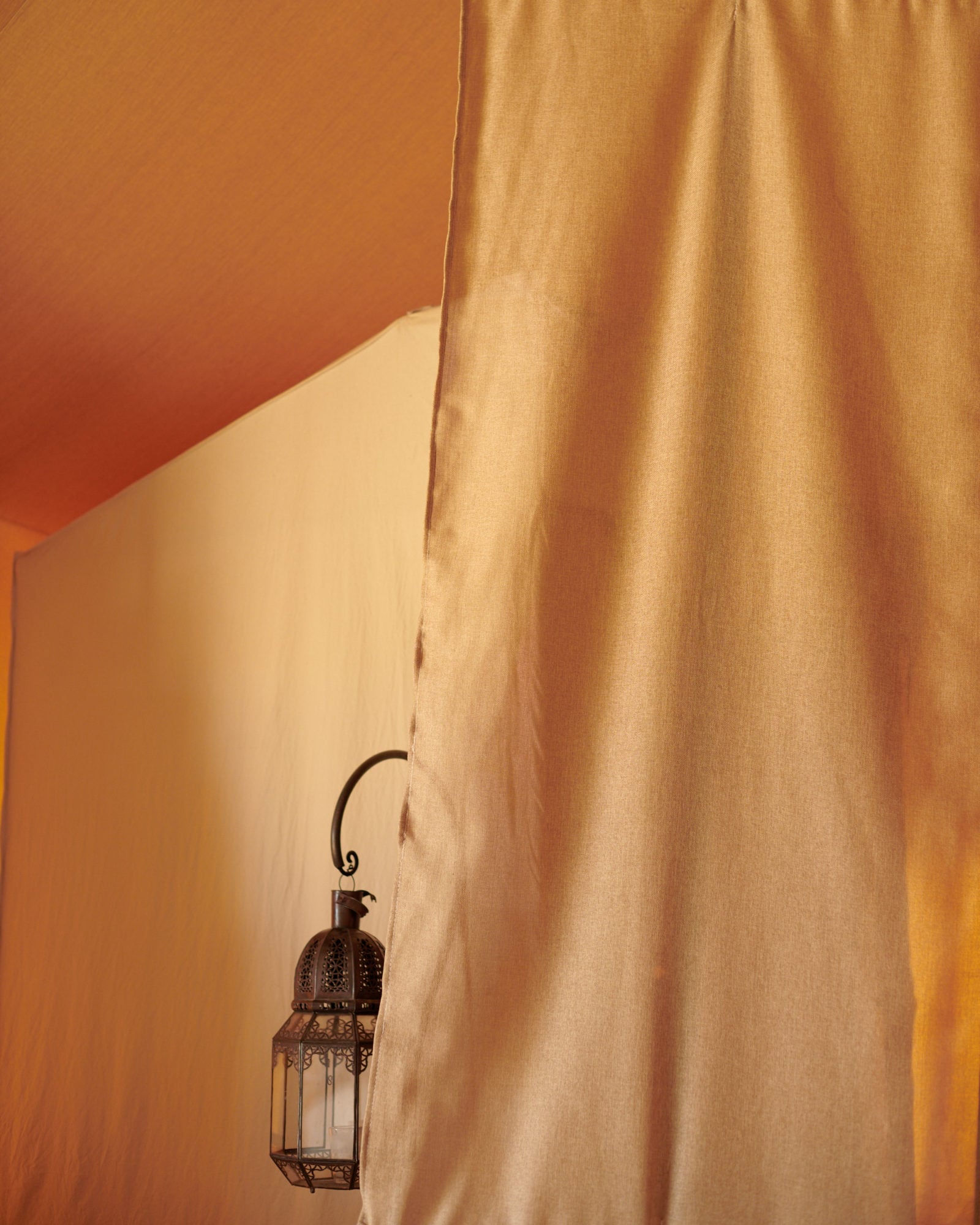 Scarabeo Camp, Africa — Ornate brass lantern suspended from ochre linen curtain against warm terracotta wall, minimalist desert aesthetic.