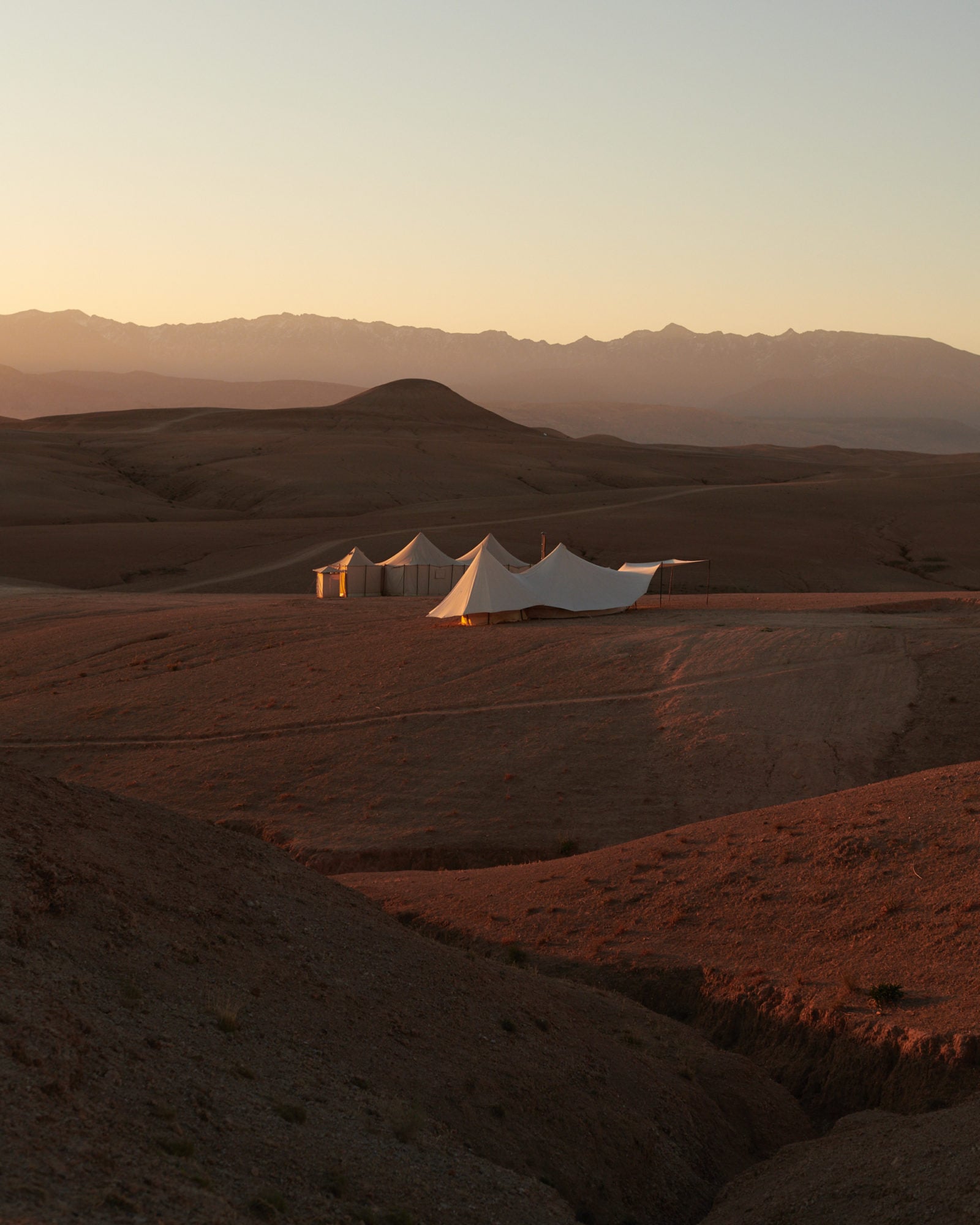 Scarabeo Camp, Africa — Luxury desert camp with white canvas tent structures at golden hour, volcanic cone and mountain range silhouetted against dusk sky.