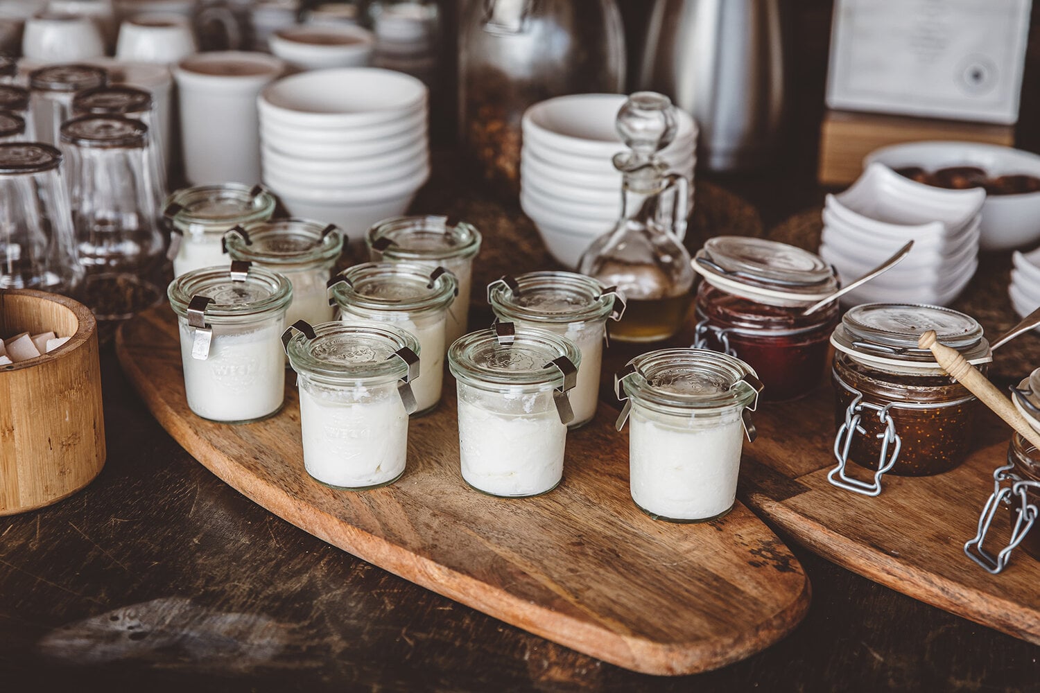 Scarabeo Camp, Africa — Glass jars with white yogurt and dark preserves arranged on wooden boards, white ceramic bowls stacked in background, warm natural light, luxury hospitality styling.