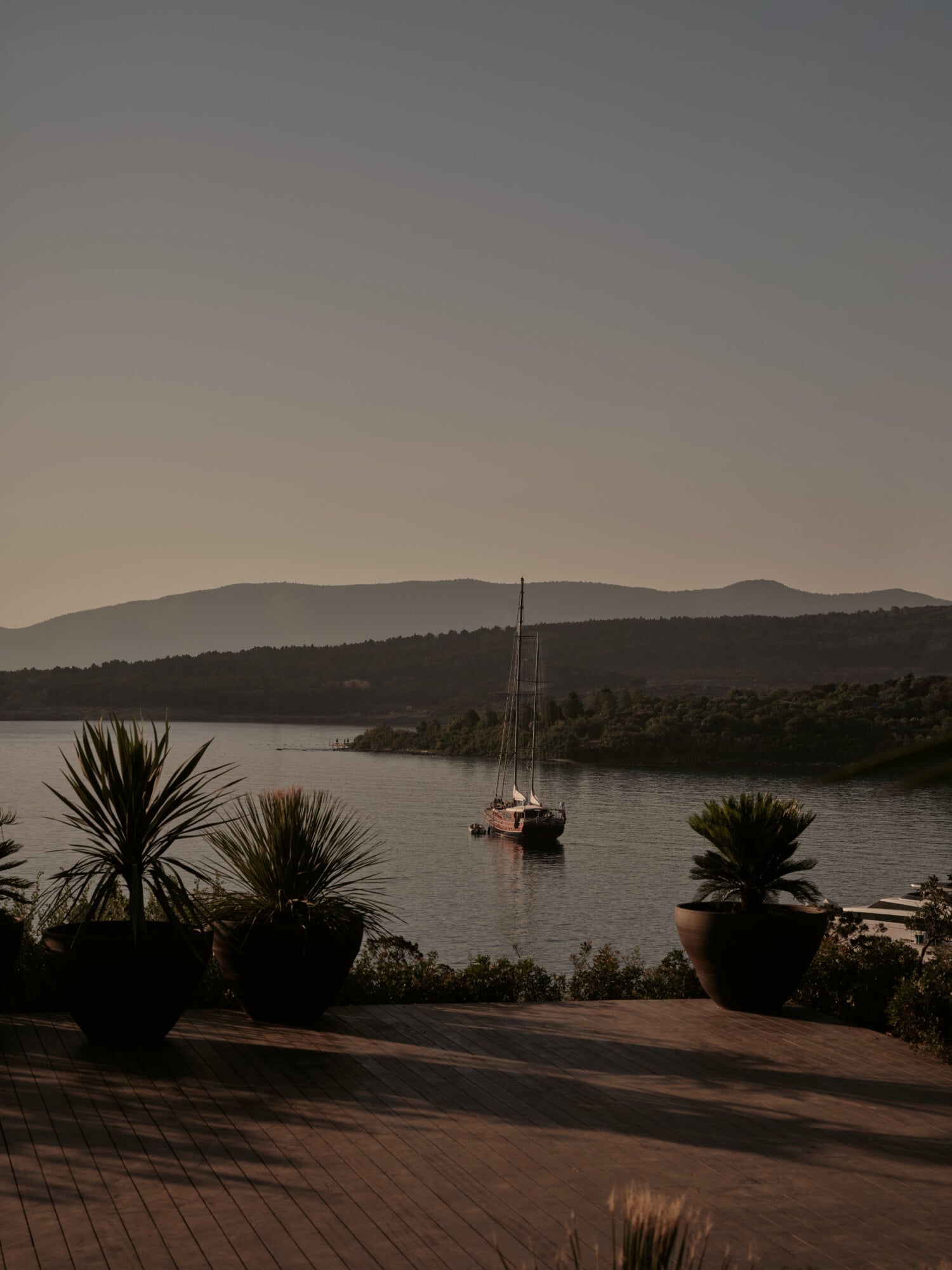 Scorpios Bodrum, Mediterranean — Sailboat anchored in calm bay at dusk with palm planters on wooden terrace, forested mountains beyond.