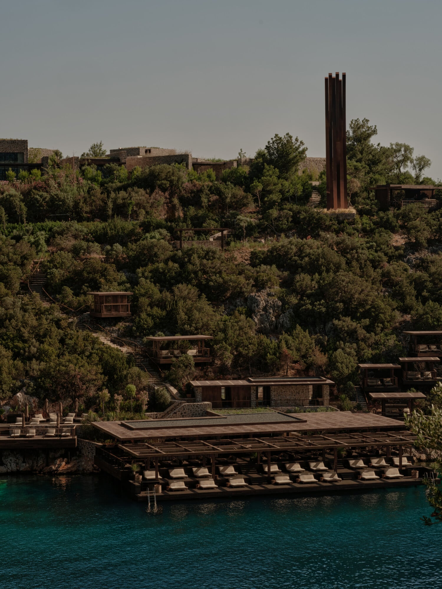 Scorpios Bodrum, Mediterranean — Terraced stone architecture with rusted metal accents overlooking turquoise water, surrounded by native vegetation on hillside.