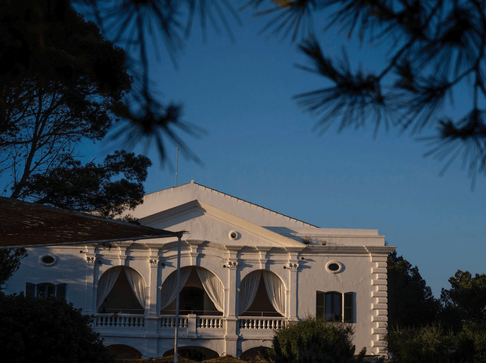 Menorca Experimental, Balearic Islands, Spain — Neoclassical villa with curved archways, Corinthian columns, and cream stone facade, framed by pine trees at golden hour.
