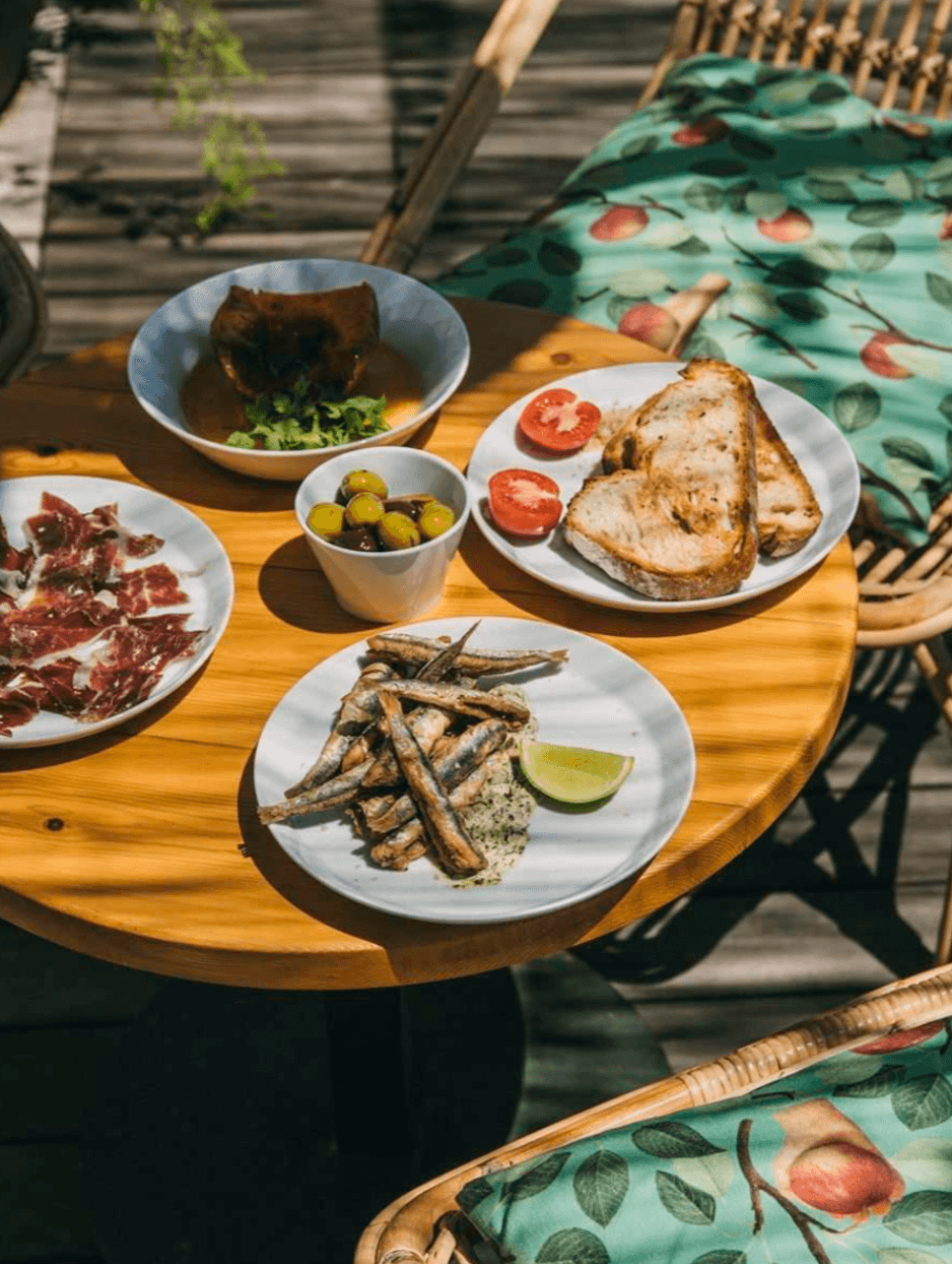 Casa Bonay, Europe — Mediterranean lunch spread with grilled fish, cured meats, olives, and bread on wooden table beneath patterned cushions.