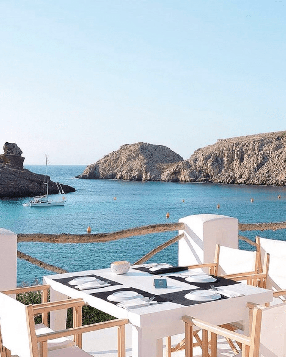 Ivette, Balearic Islands — Seaside terrace dining overlooking turquoise cove with limestone cliffs, white chairs, natural wood accents, sailboat anchorage.