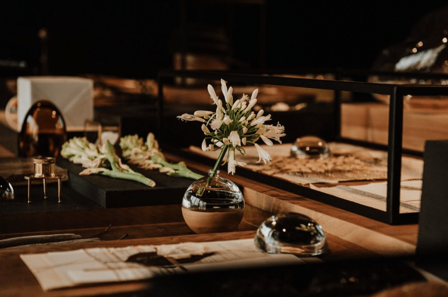 Xinú, Mexico — Minimalist wooden desk with white agapanthus in spherical vase, brass accents, and desert landscape visible through black-framed windows at dusk.