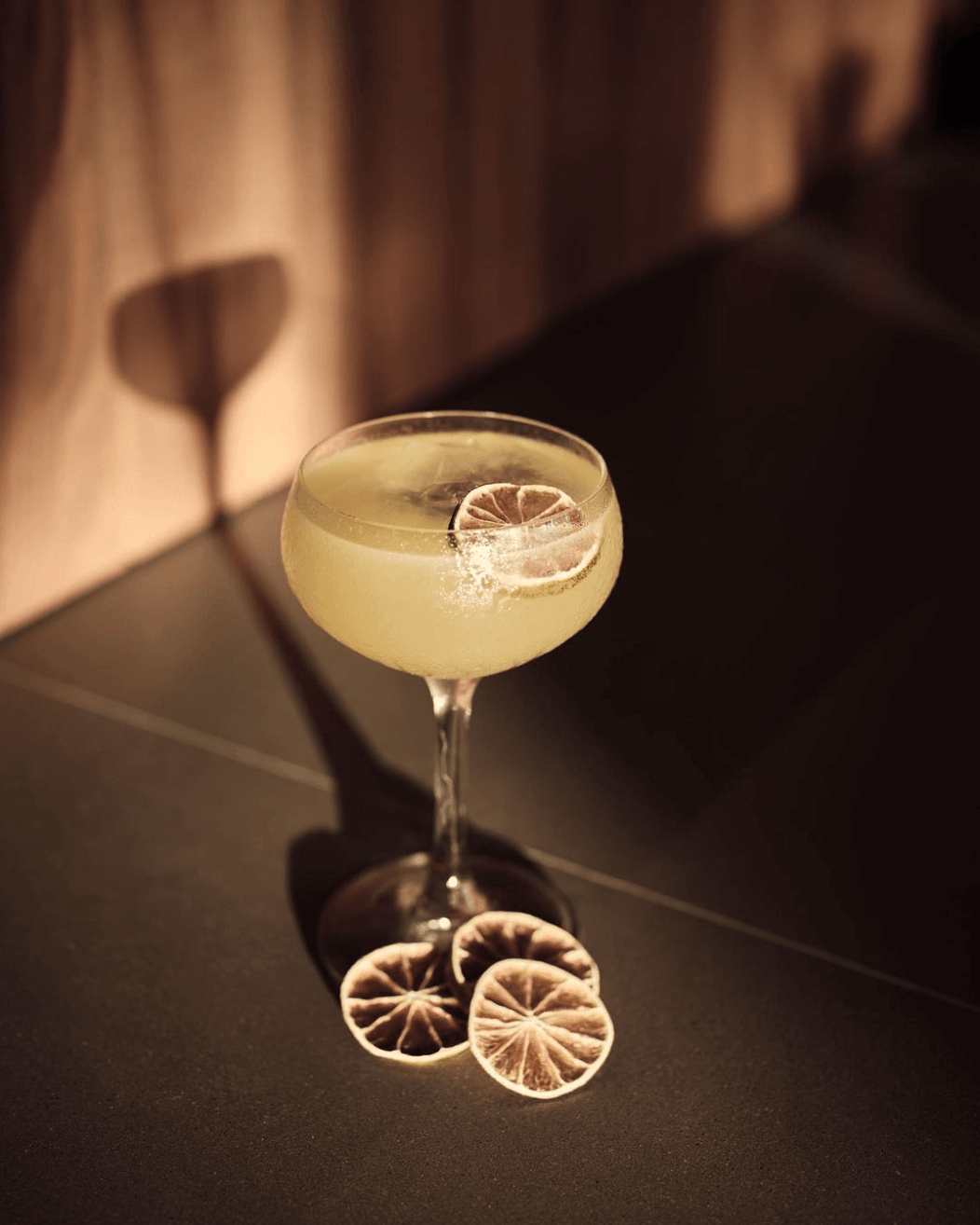 Fari Beach Club, Asia — Pale citrus cocktail with lemon slice garnish on dark surface, casting geometric shadows in warm afternoon light.