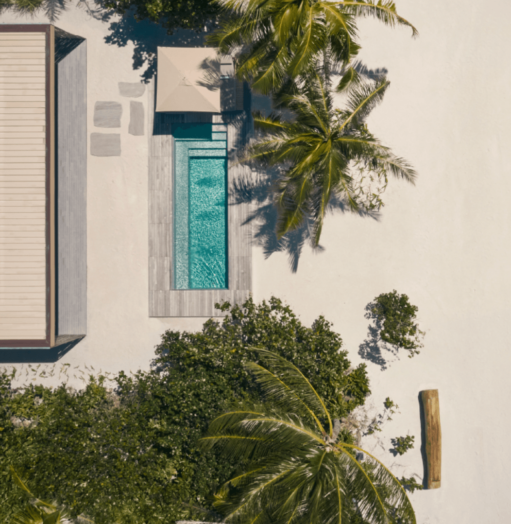 Patina Maldives, Asia — Aerial view of minimalist beachfront villa with turquoise pool, white walls, palm trees and geometric shadows in tropical luxury resort setting.