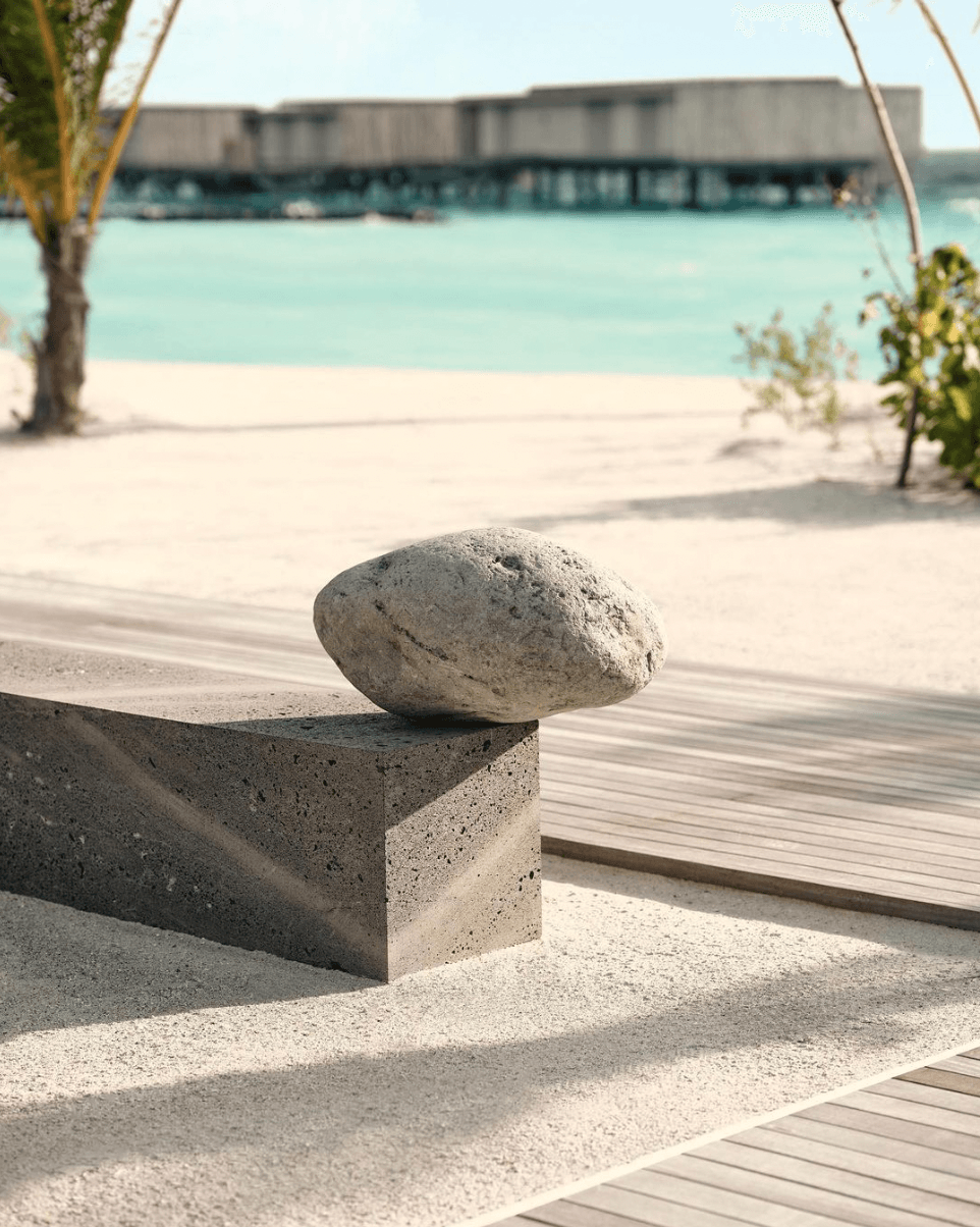 Patina Maldives, Maldives — Concrete sculptural element with smooth stone on turquoise lagoon beach, modern minimalist resort architecture and wooden deck.