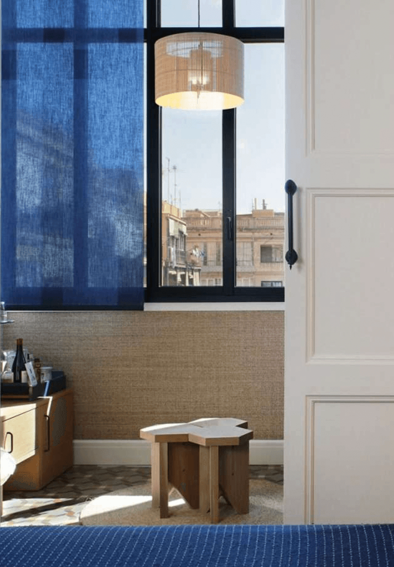 Casa Bonay, Europe — Modern hotel room with natural light, woven wall paneling, wooden furniture, blue textiles, and black-framed window overlooking classical architecture.