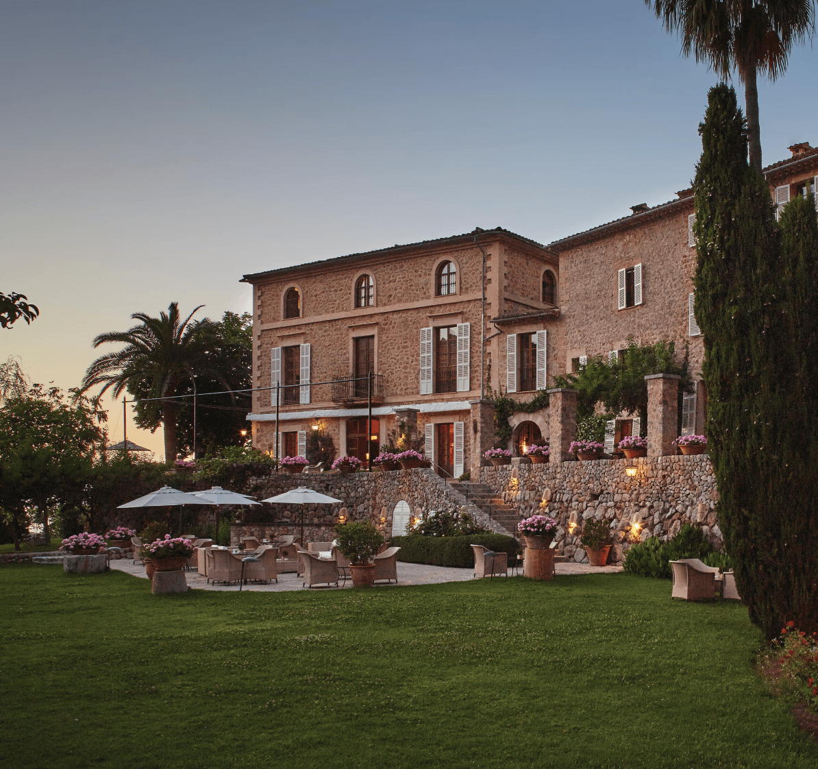 Belmond La Residencia, Balearic Islands — Tuscan stone villa at dusk with manicured lawn, terrace seating, purple hydrangeas, cypress trees, and warm architectural lighting.