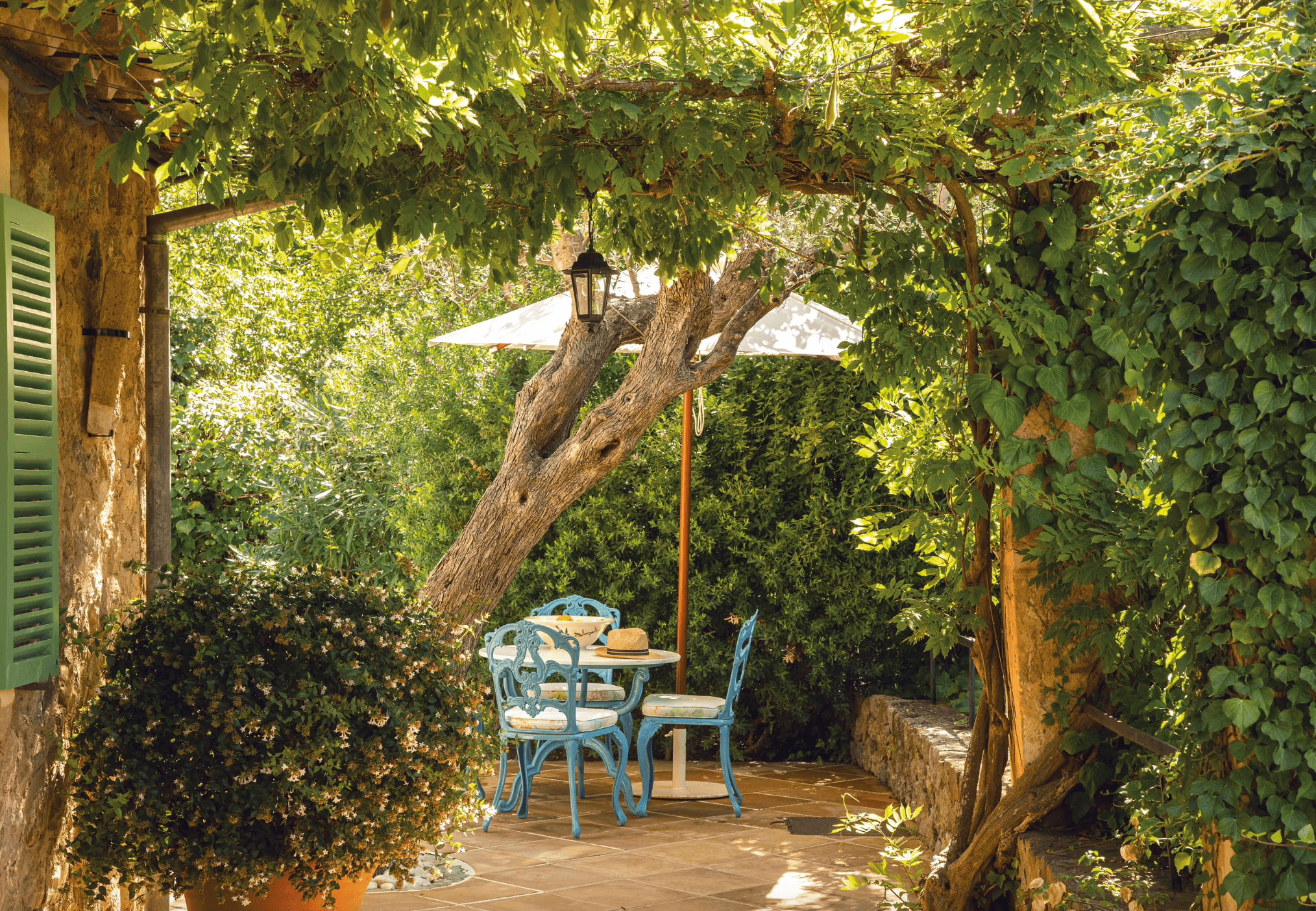 Belmond La Residencia, Balearic Islands, Spain — Blue bistro chairs beneath ancient tree with ivy-draped stone walls and dappled sunlight in Mediterranean garden courtyard.