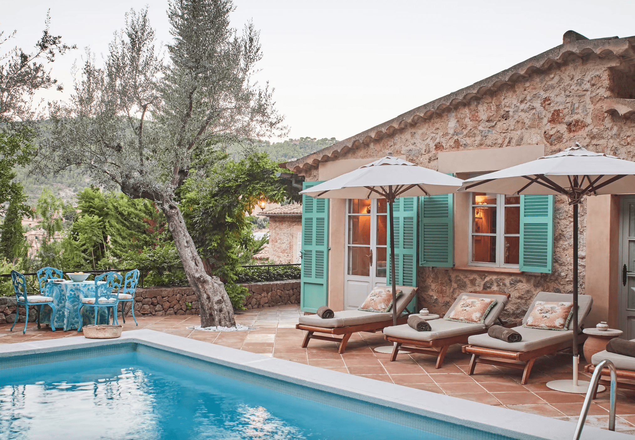 Belmond La Residencia, Balearic Islands, Spain — Stone villa with turquoise shutters, poolside loungers under umbrellas, ancient olive tree, Mediterranean terracotta courtyard.