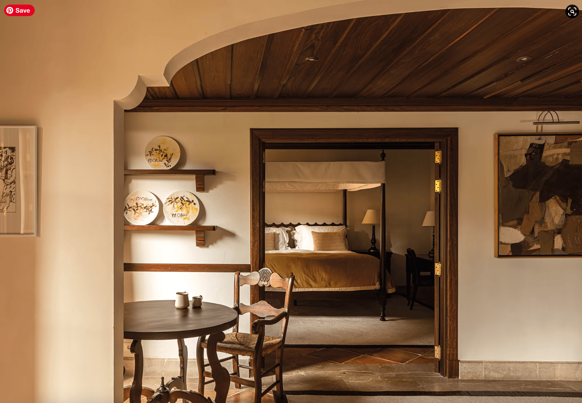 Belmond La Residencia, Balearic Islands — Warm-toned suite with dark wood ceiling, round dining table, decorative ceramic plates, and canopied bed visible through open doorway.