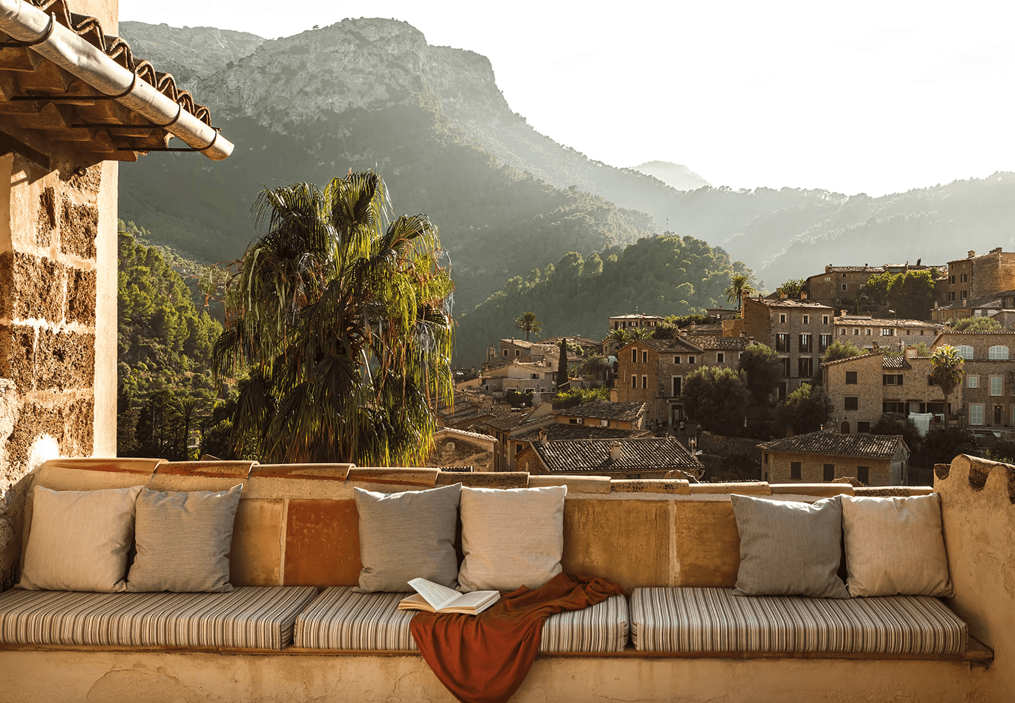 Belmond La Residencia, Balearic Islands, Spain — Striped daybed with cushions on stone terrace overlooking palm tree, village rooftops, and forested mountains in golden afternoon light.