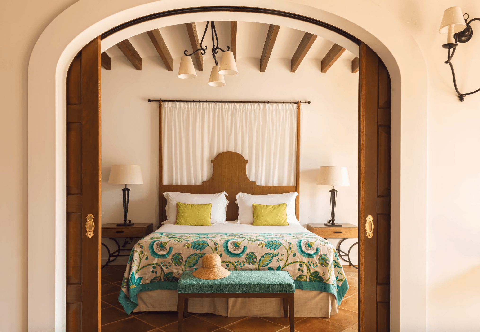 Belmond La Residencia, Balearic Islands — Arched bedroom alcove with wooden beams, canopy bed, turquoise patterned bedspread, cream curtains, pendant lights, and terracotta floors.