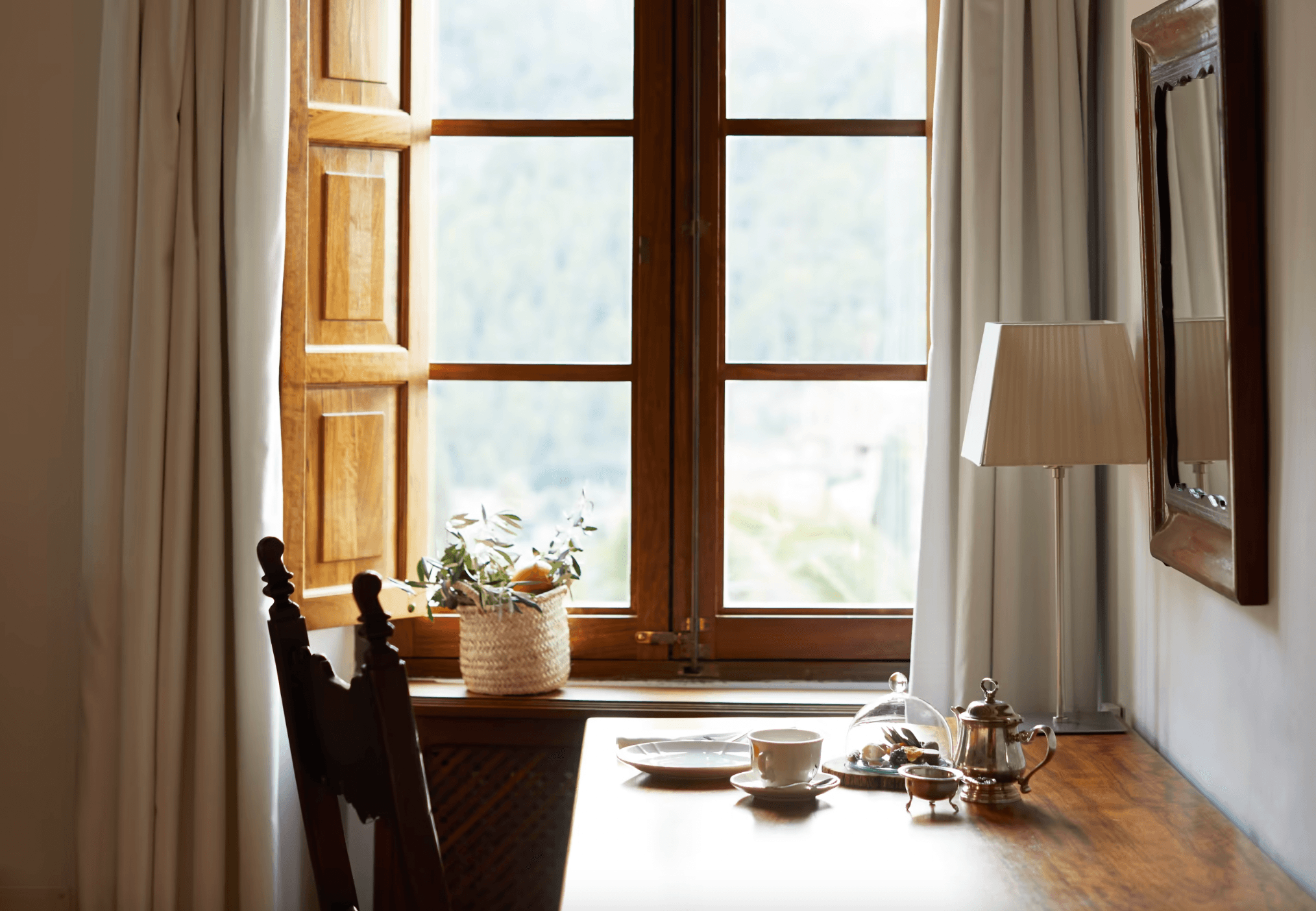 Belmond La Residencia, Balearic Islands, Spain — Sunlit dining room with wooden-framed windows, cream curtains, woven basket with greenery, tea service, and dark wooden chair overlooking mountain landscape.