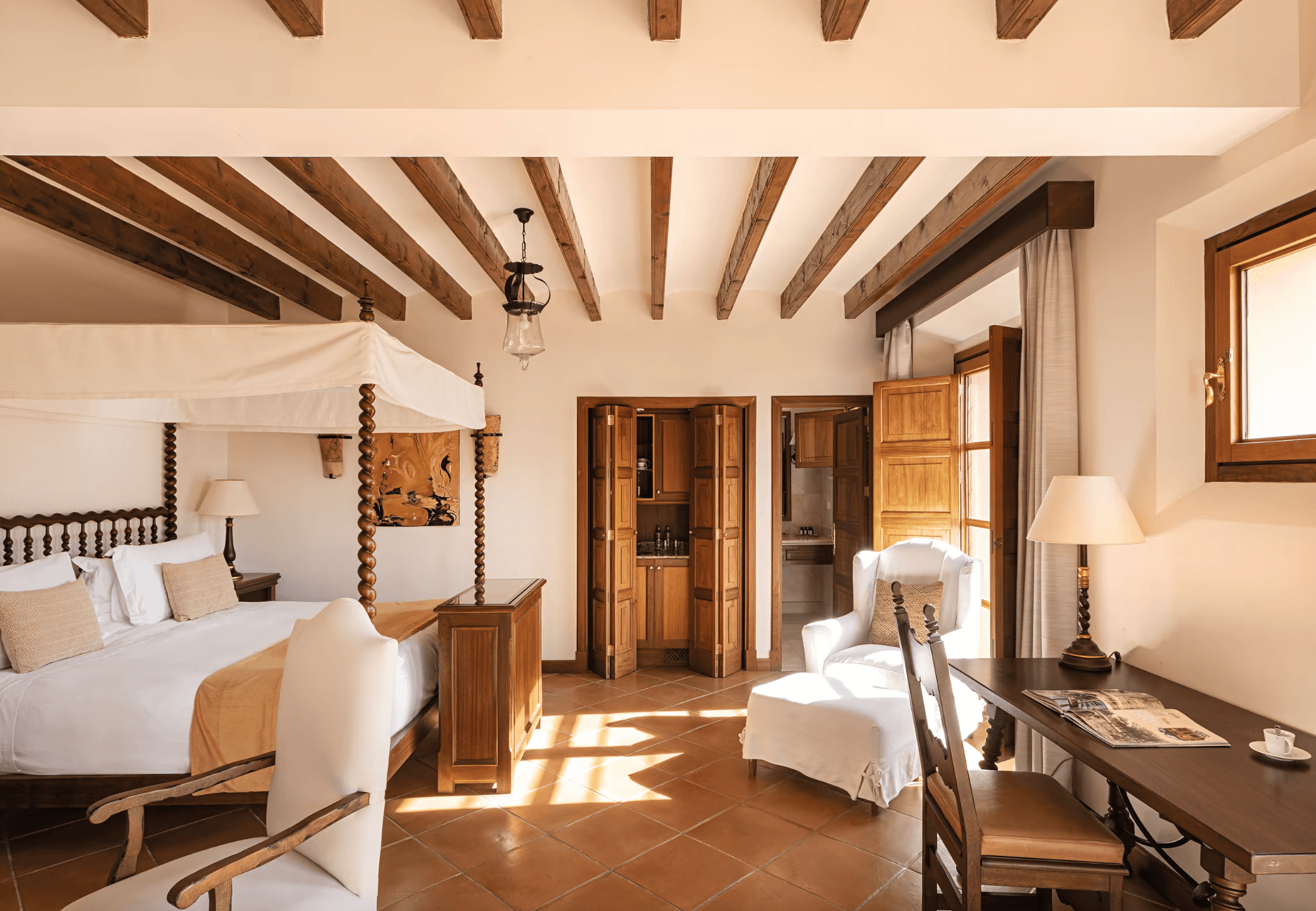 Belmond La Residencia, Balearic Islands — Spacious bedroom with exposed wooden beams, canopy bed, terracotta floors, and arched doorways opening to Mediterranean light.