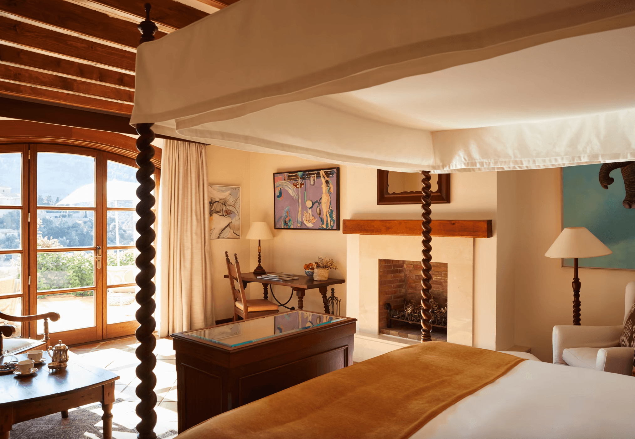 Belmond La Residencia, Balearic Islands — Luxury suite with canopied bed, wooden furnishings, fireplace, and Mediterranean terrace views through French doors.