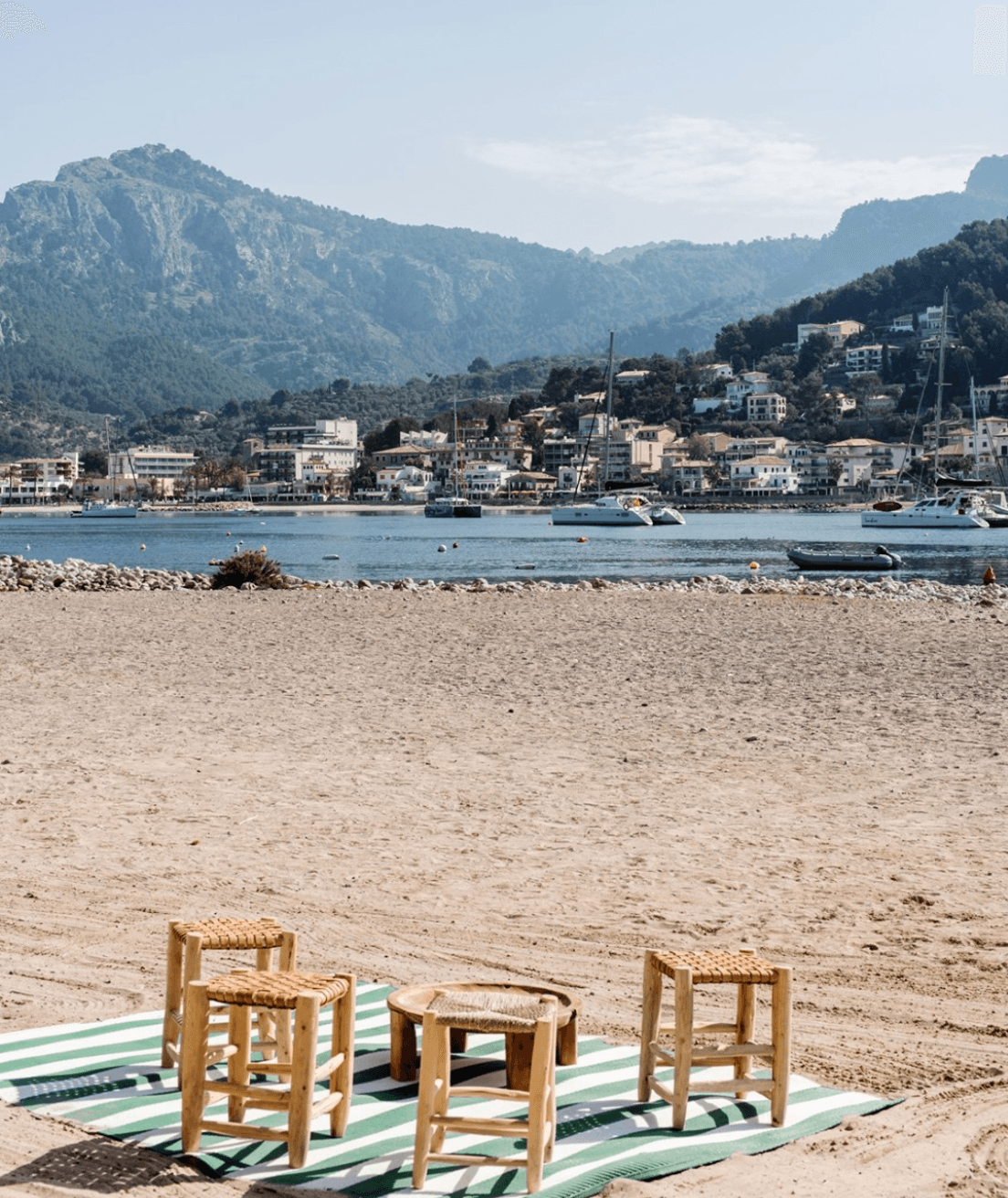 Wooden stools on striped linen at Patiki Beach, turquoise water and limestone peaks framing a Mediterranean harbor town.