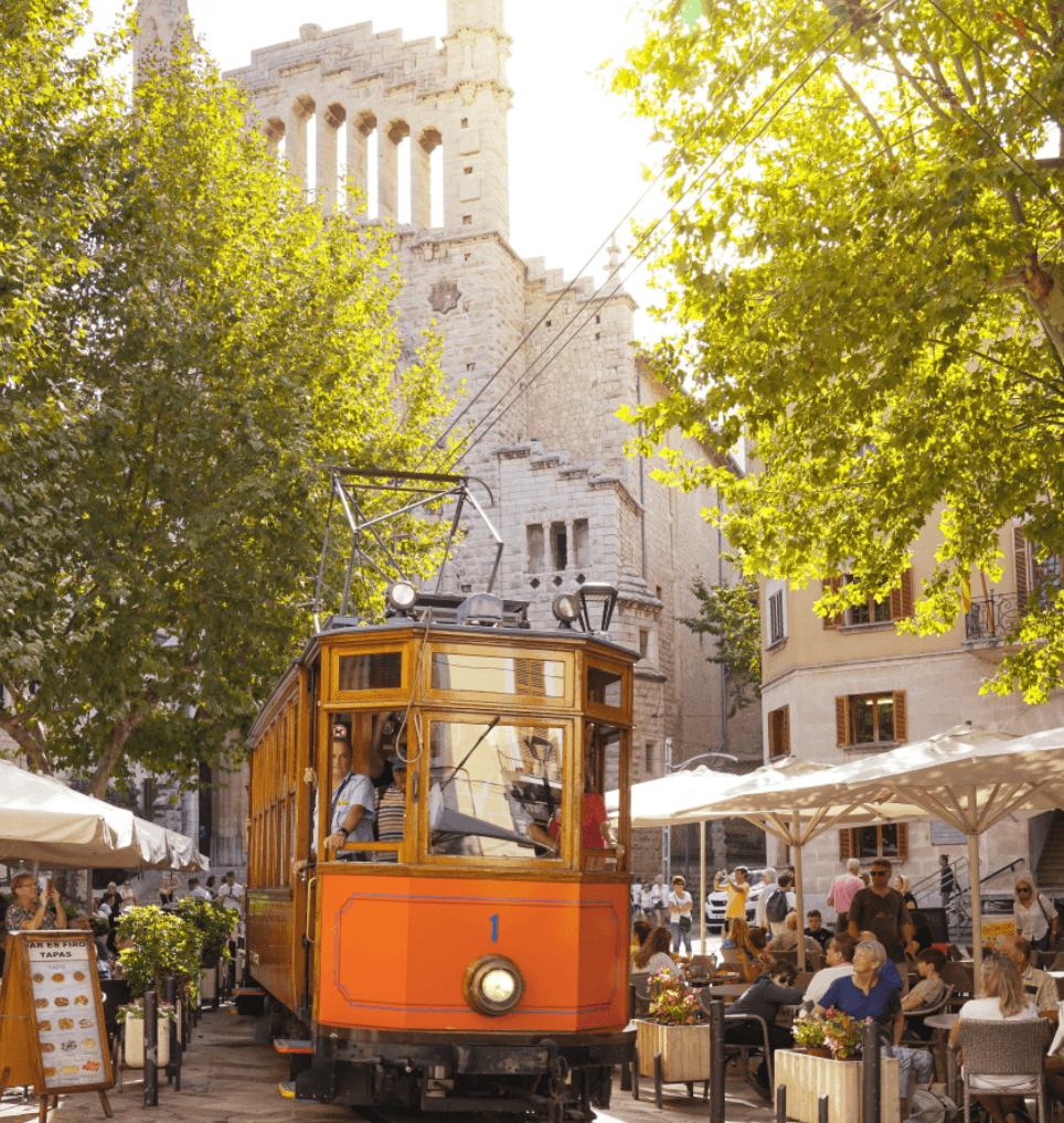 Soller, Balearic Islands — Historic orange tram on tree-lined plaza with stone tower, café seating, and dappled sunlight.