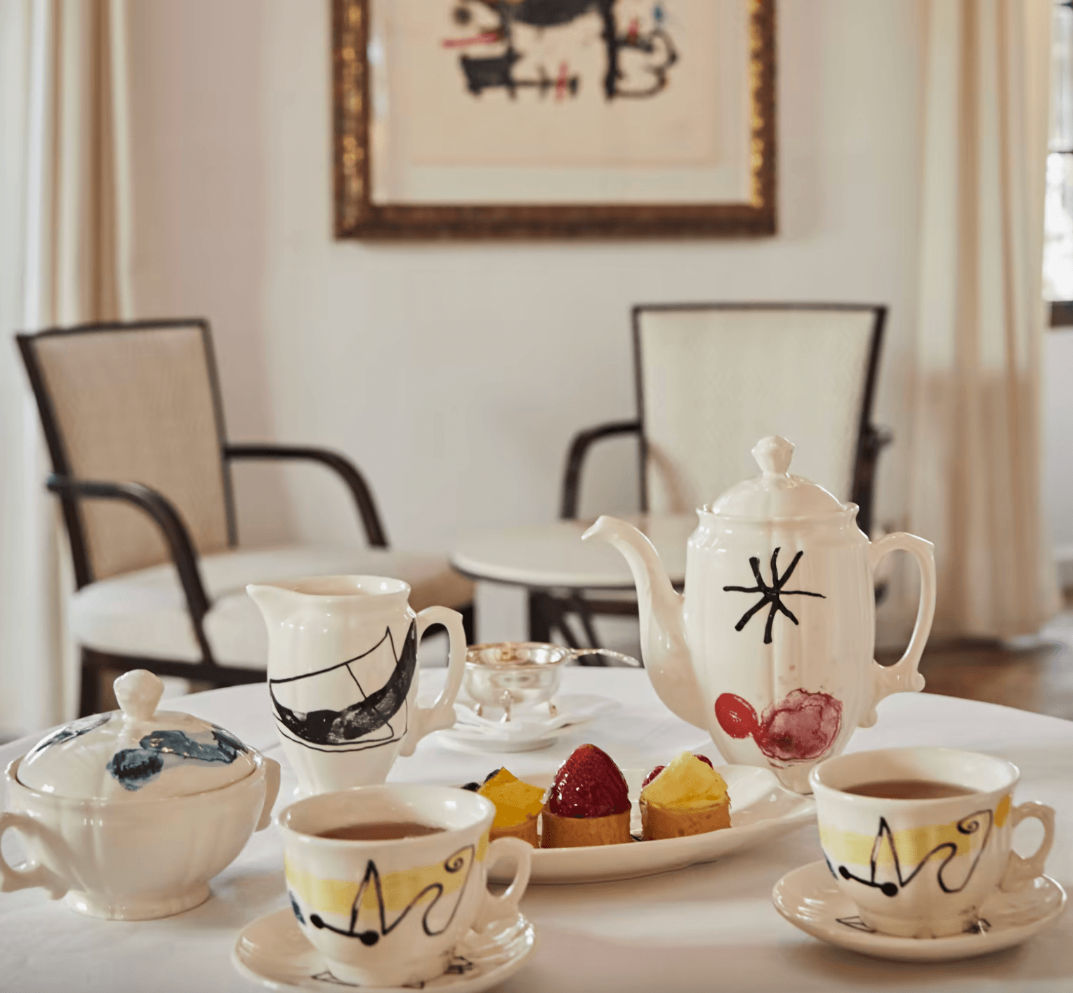 Tea with Miro, Balearic Islands, Spain — Hand-painted ceramic teapot and cups with modernist line drawings, pastries on white plate, minimalist interior with lounge chairs and artwork.
