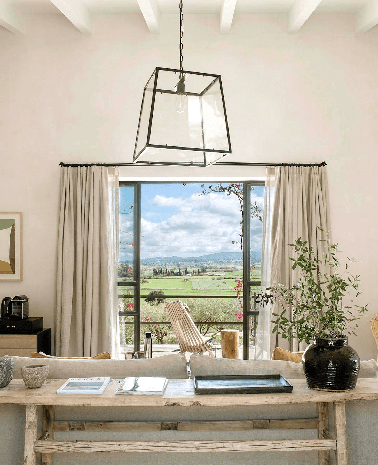 Finca Serena, Balearic Islands, Spain — Minimalist interior with stone desk, metal pendant light, and panoramic countryside views through floor-to-ceiling doors with linen curtains.