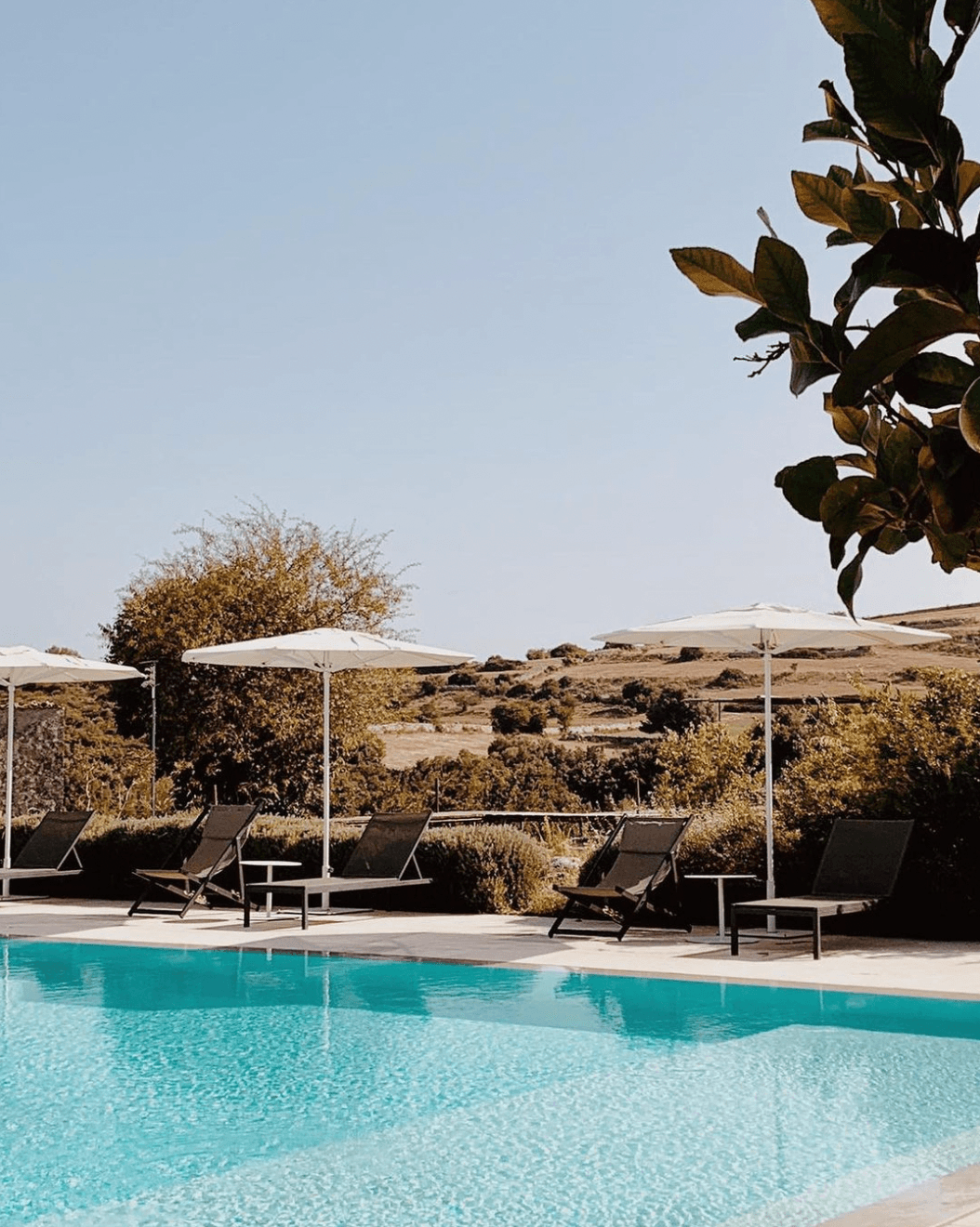 Dimora delle Balze, Europe — Infinity pool with turquoise water, black loungers, white umbrellas, and rolling countryside landscape under clear sky.