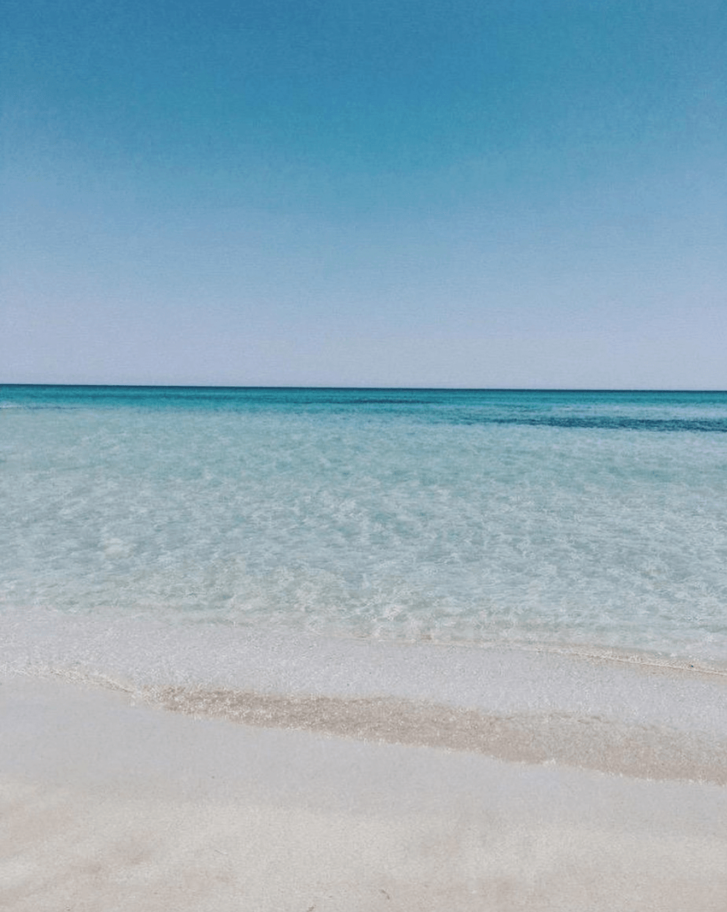 Crystalline turquoise water meets pale sand under clear sky at Fontane Bianche beach.