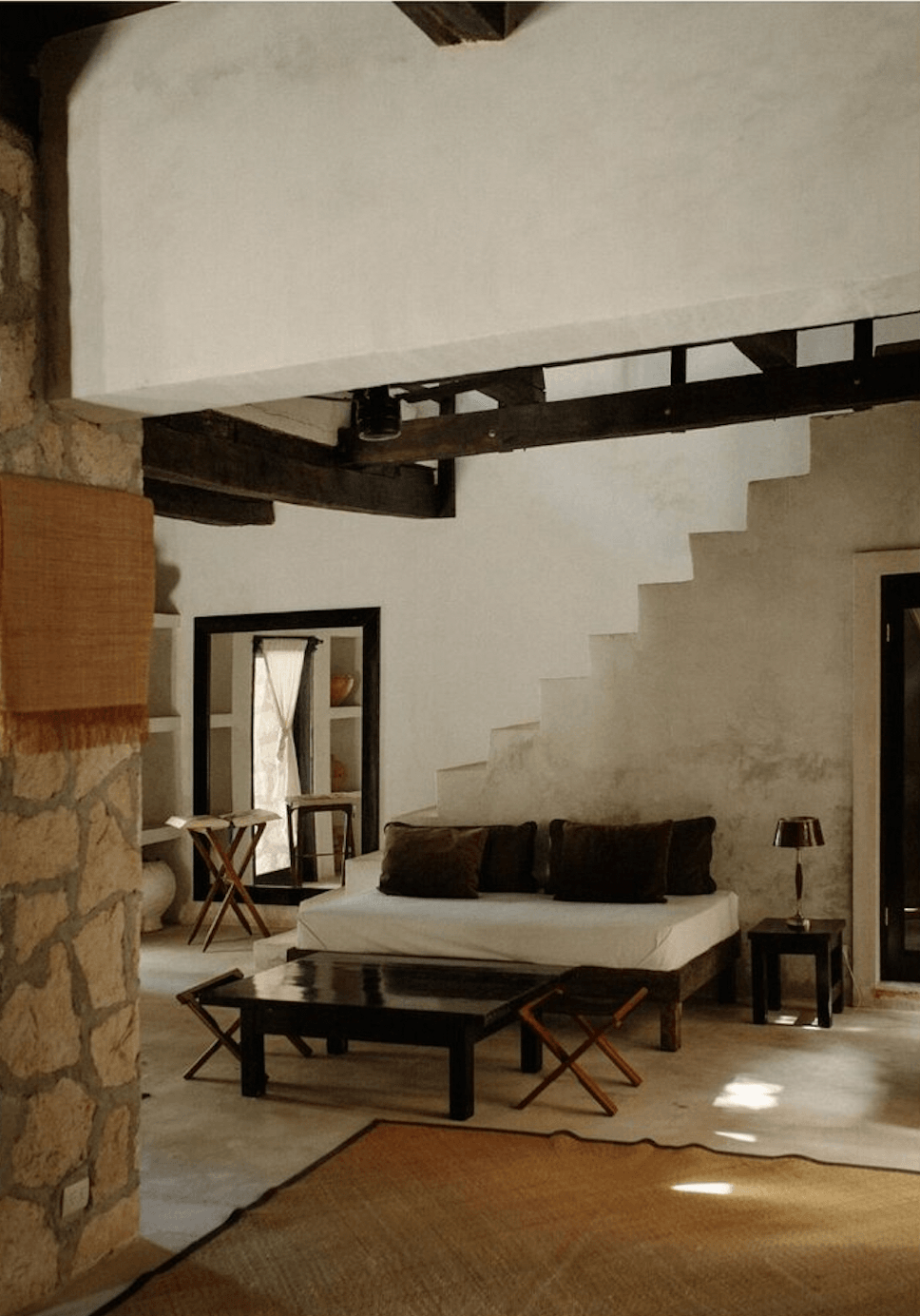 Coqui Coqui Papholchac, Mexico — Minimalist bedroom with whitewashed walls, dark wood beam, stepped architectural details, stone accents, and natural light filtering through open doorway.