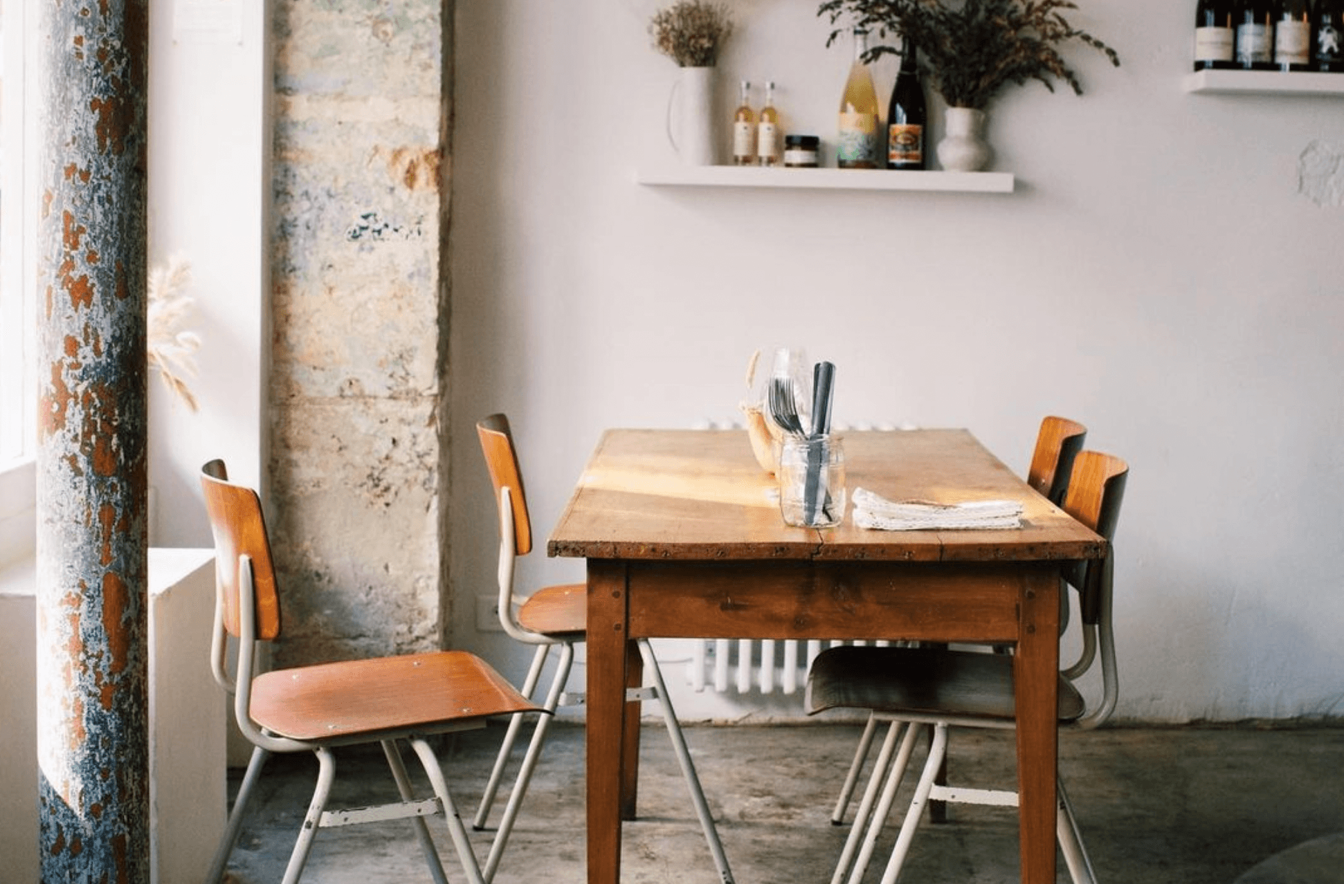 Early June, Europe — Minimalist dining space with weathered concrete column, wooden table, bentwood chairs, industrial metal frame, white walls, open shelving with wine bottles, natural light, contemporary hospitality design.