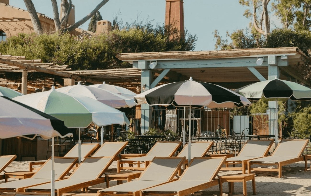 A sun-drenched beach club terrace with wooden loungers, sage and cream umbrellas, and rustic timber structures beneath clear sky.