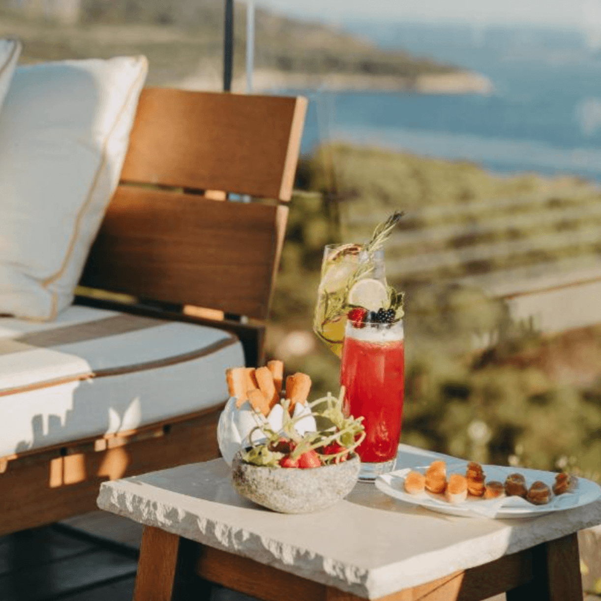 Lily of the Valley, Europe — Coastal terrace with aperitif cocktail, fresh fruit bowl, pastries, and wooden loungers overlooking Mediterranean landscape.