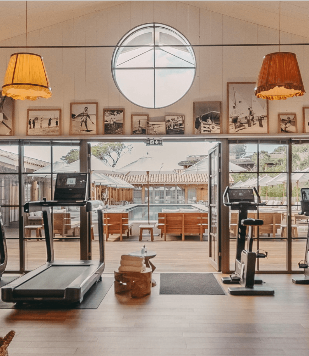 The Shape Club — Shape Club fitness studio with wooden beams, pendant lamps, floor-to-ceiling glass doors opening to courtyard, treadmills and exercise bikes, sepia-toned surf photography, circular skylight, minimalist luxury wellness design.