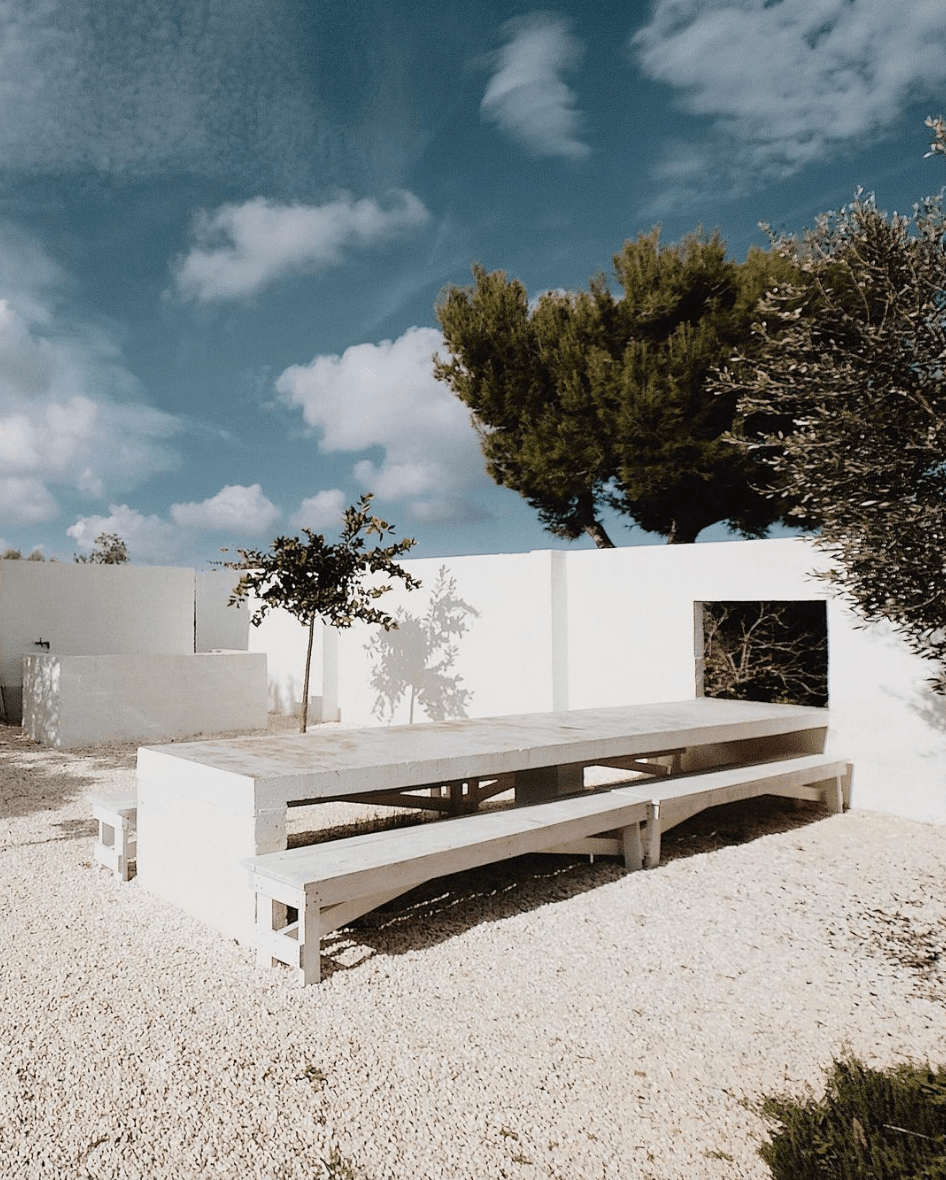 Masseria Moroseta, Europe — Minimalist whitewashed concrete table and benches in gravel courtyard with cypress trees and blue sky, Mediterranean architecture detail.
