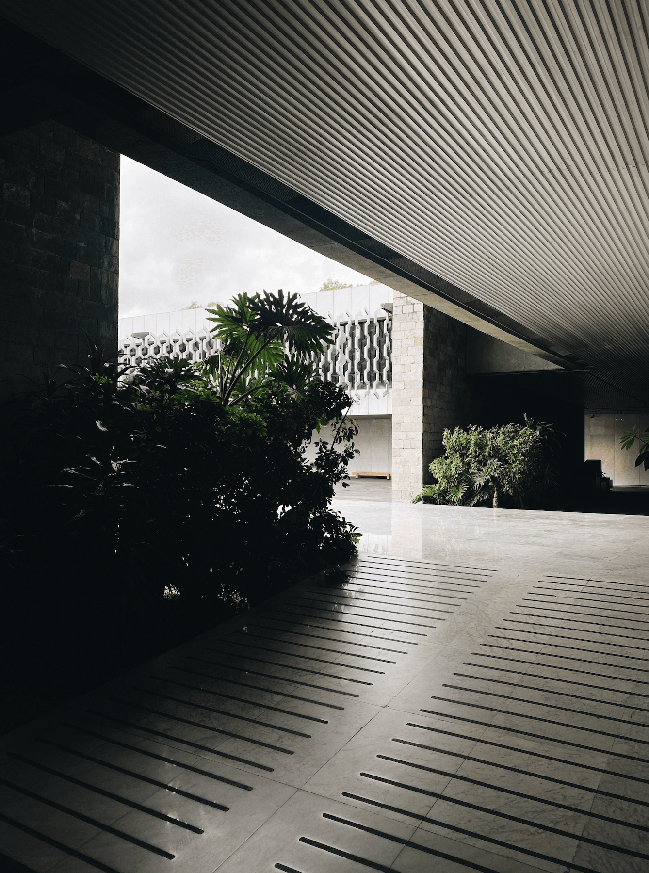 Museo Nacional de Antropología, Mexico City, Mexico — Modernist courtyard with corrugated metal canopy, stone facade, lush tropical plants, overcast daylight filtering through geometric architecture.