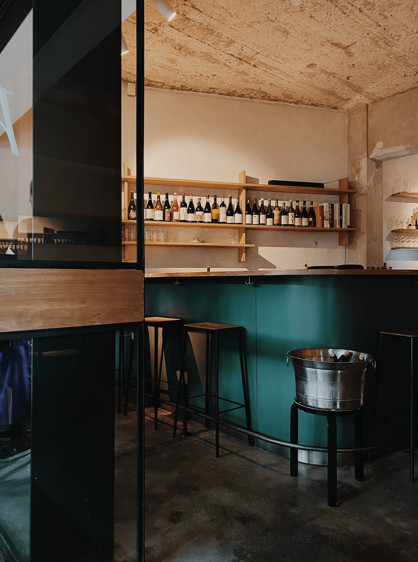 Hugo Wine Bar, Mexico — Minimalist wine bar with teal-painted walls, natural wood shelving displaying bottles, black bar seating, concrete floors, and industrial metal ice bucket.