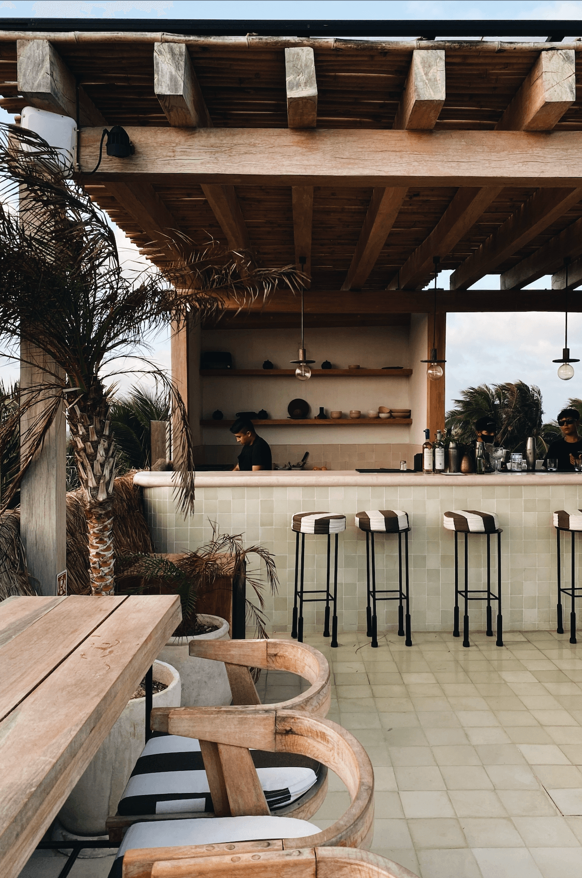 RoOf Sunset Bar, Mexico — Outdoor bar counter with striped stools, wooden pergola, palm plants, and cream tile flooring overlooking coastal views.