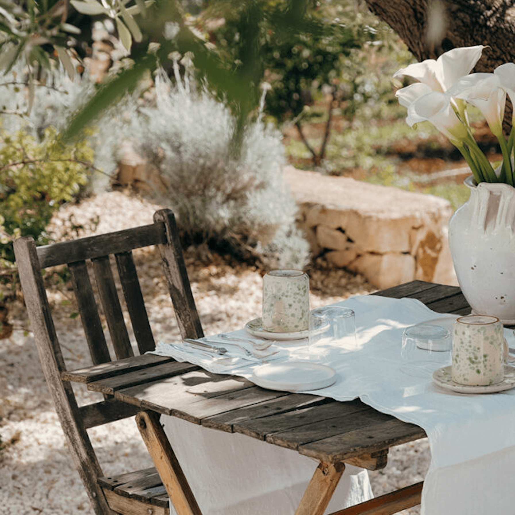 Masseria Moroseta, Europe — Rustic wooden table with white linens set in Mediterranean garden, white flowers and stone architecture, dappled sunlight.