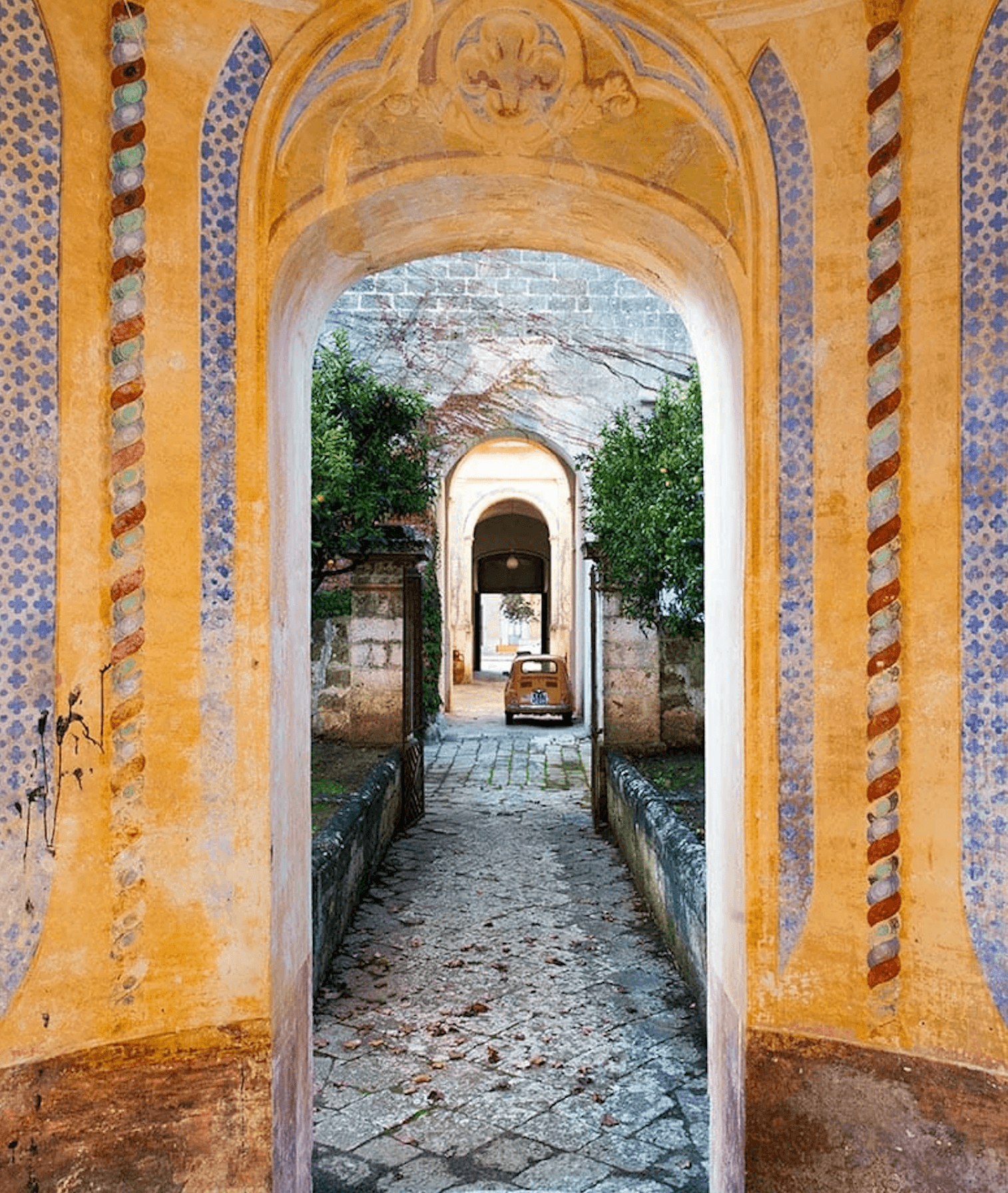Palazzo Daniele, Europe — Arched passageway with ochre walls, blue-patterned tilework, and ivy-draped stone courtyard leading through successive archways.