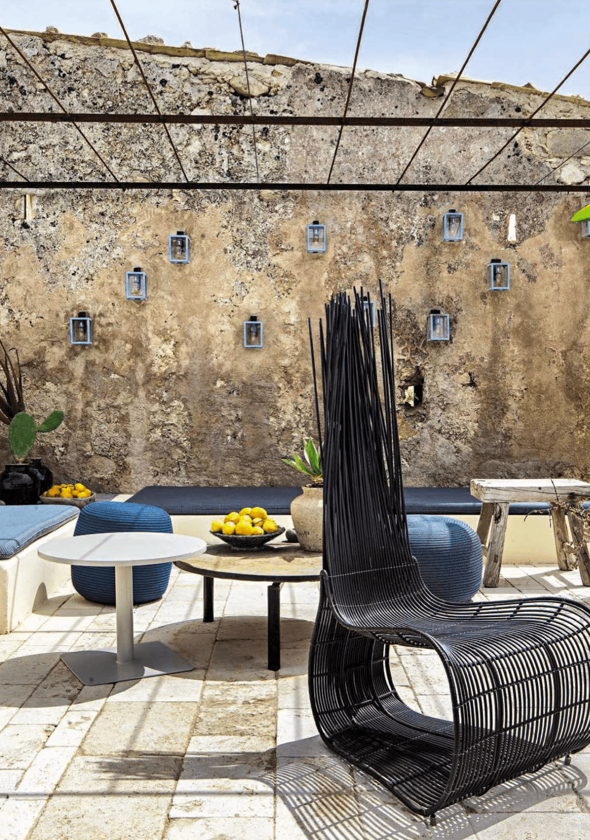 Dimora delle Balze, Europe — Weathered stone courtyard with black woven lounge chair, white round table, potted citrus, and blue cushioned seating beneath pergola.