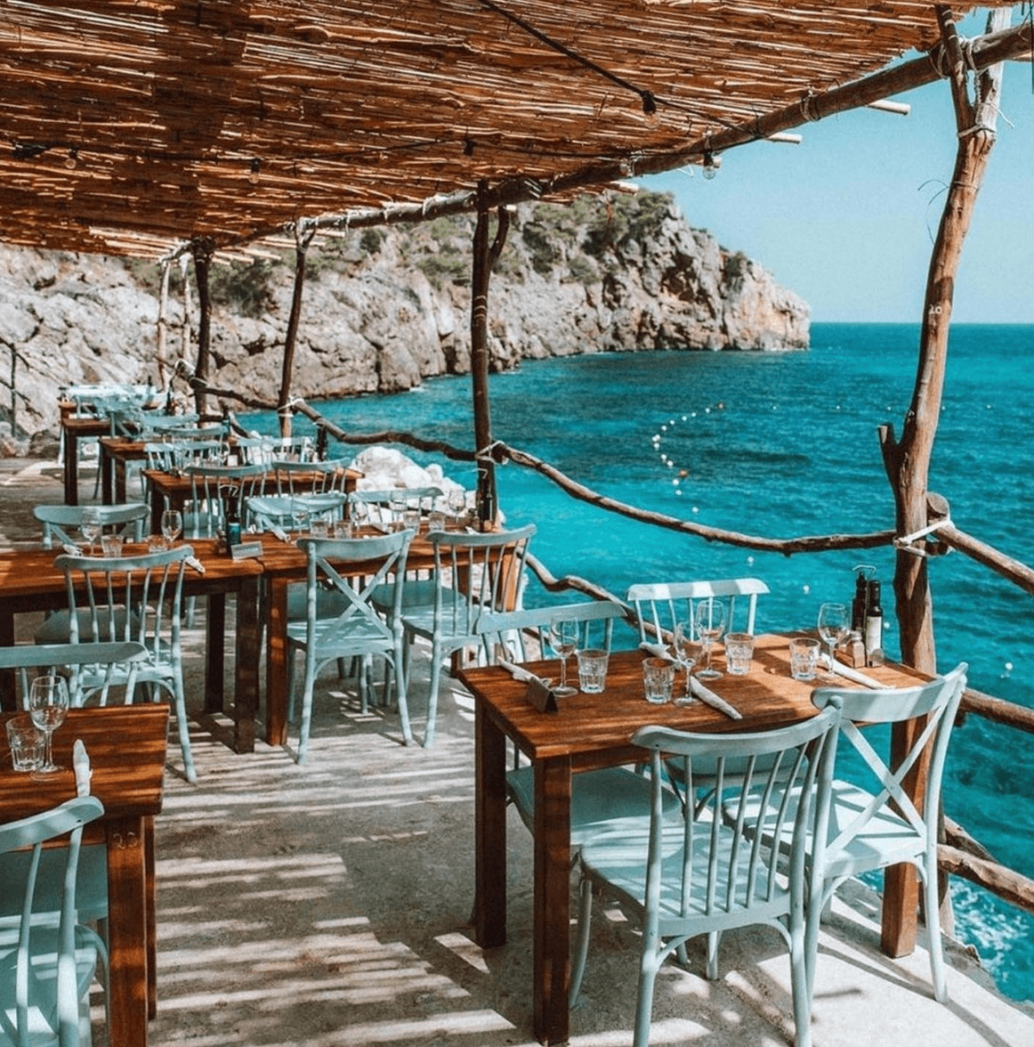 Ca's Patró March, Balearic Islands, Spain — Seaside restaurant terrace with wooden tables, pale blue chairs, driftwood pergola, turquoise Mediterranean waters and limestone cliffs.