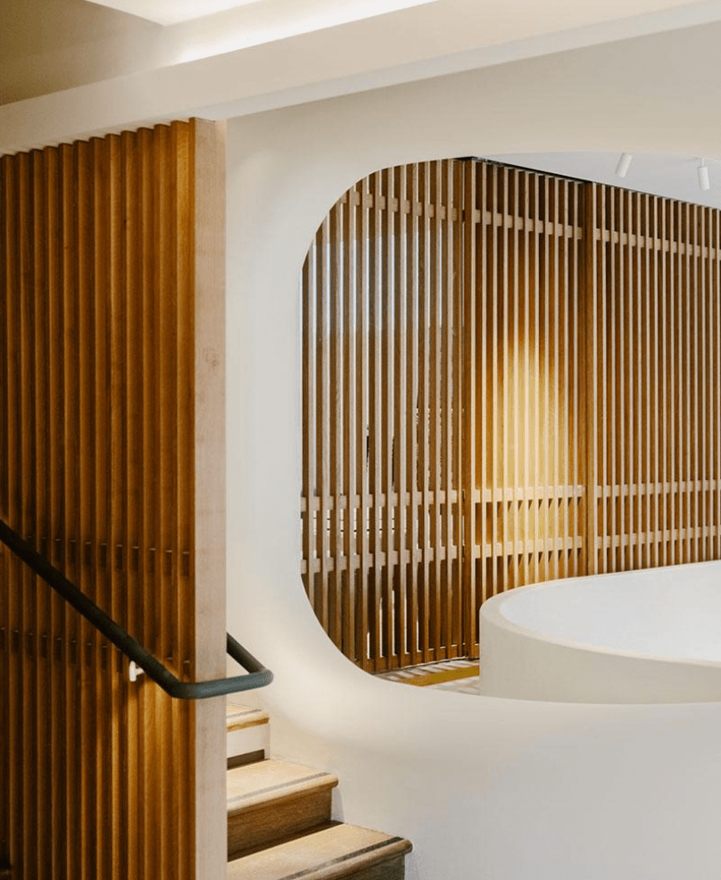 Belle Plage, Europe — Curved wooden slat partition with arched opening beside white sculptural counter and floating staircase in minimalist luxury interior.