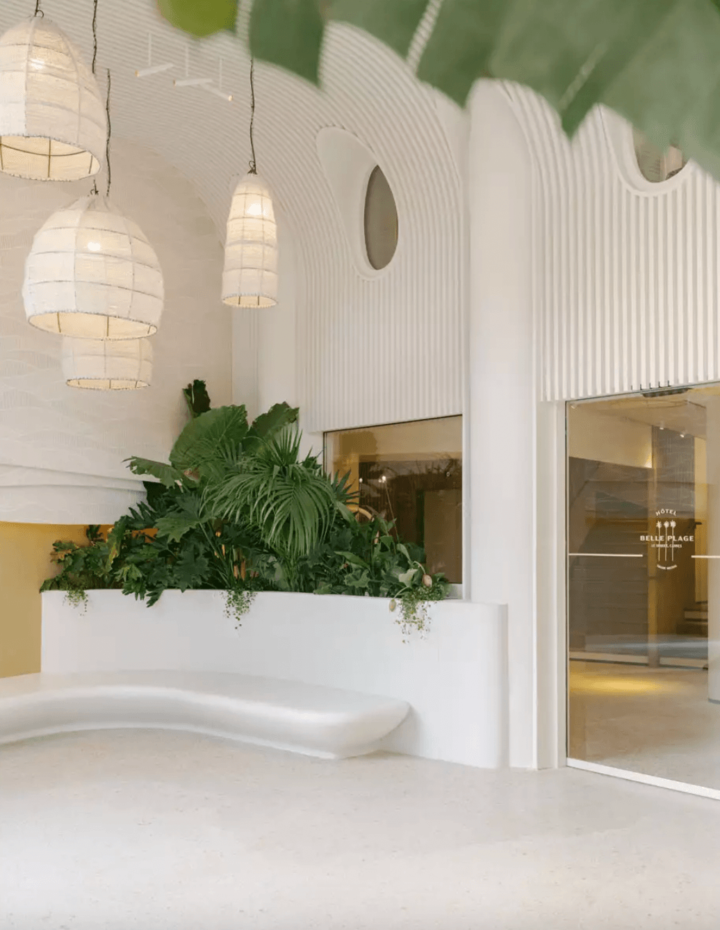 Belle Plage, Europe — Minimalist white lobby with curved architectural details, pendant lanterns, lush planter, and golden accent wall creating serene luxury hospitality aesthetic.