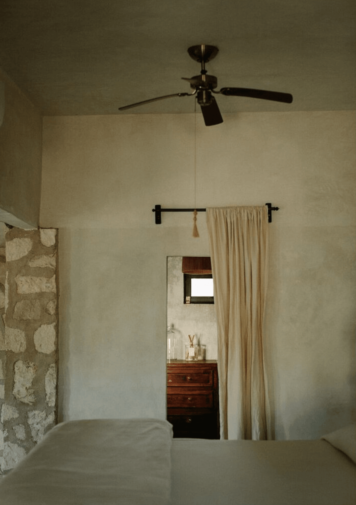 Coqui Coqui Papholchac, Mexico — Minimalist bedroom with cream-draped doorway, black ceiling fan, wooden dresser, and textured stone accent wall.