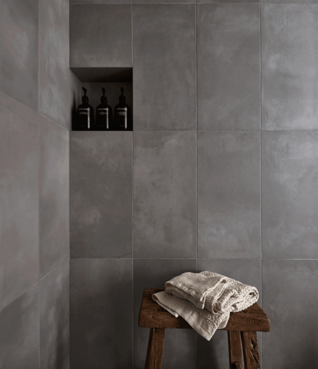 The Surfrider, North America — Minimalist bathroom niche with three black bottles on concrete tile wall, natural linens folded on rustic wooden stool, spa aesthetic.